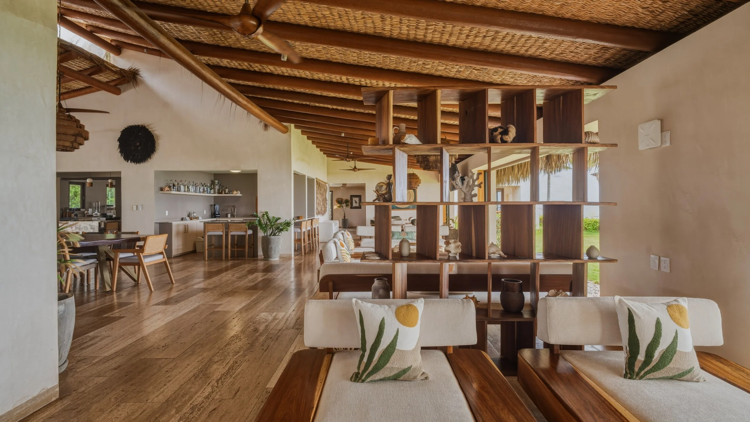 Interior of a tropical-style living room featuring a wooden ceiling, a wooden room divider with shelves holding decorative items, and a view of the outdoor greenery through large windows.