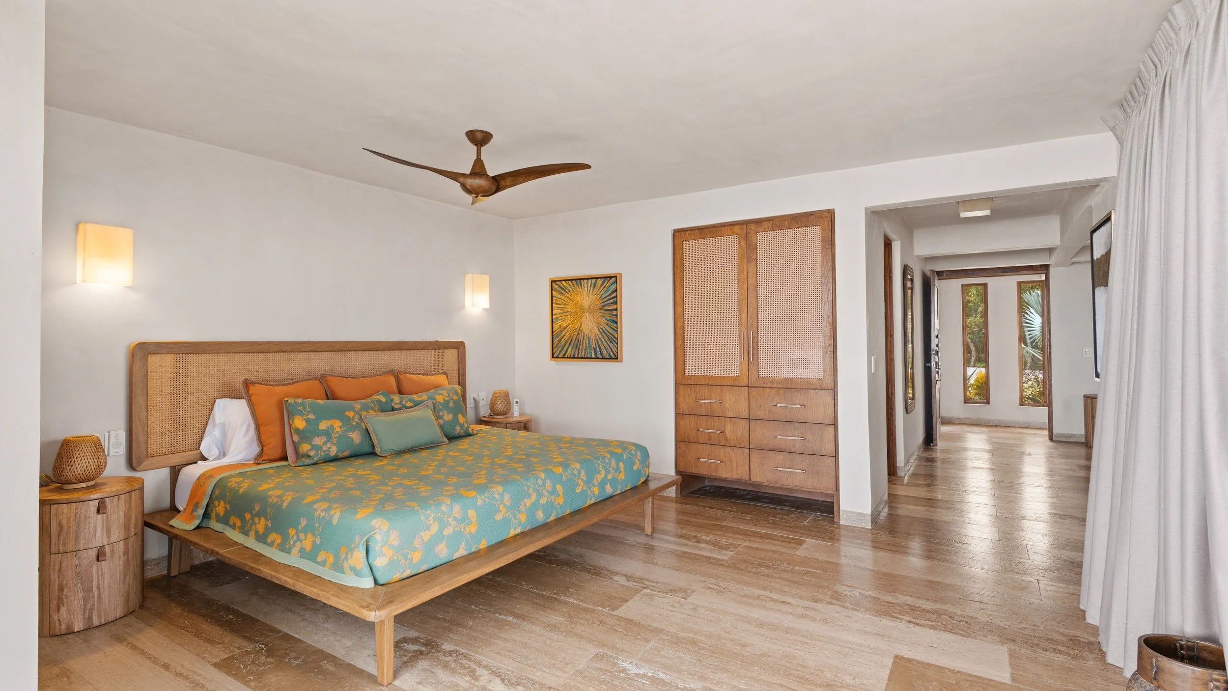 A bedroom with a wooden bed frame and colorful bedding, wooden nightstands, and built-in wooden closet, with hardwood floors, white walls, and natural light coming through windows with curtains.