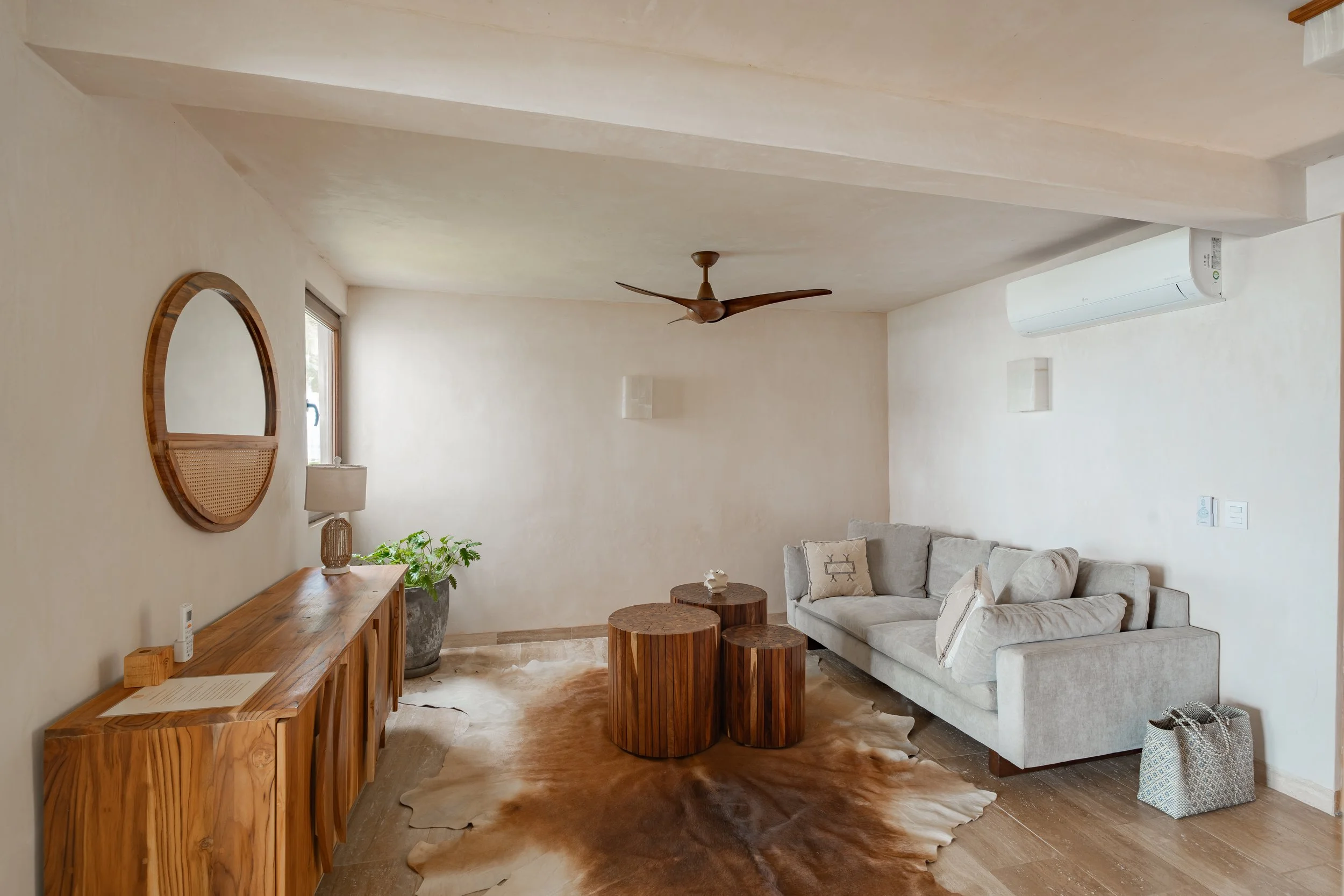 Living room with a beige sofa, wooden coffee tables, a wooden sideboard, a round mirror on the wall, a white air conditioning unit, a ceiling fan, a window, and a cowhide rug on the wooden floor.