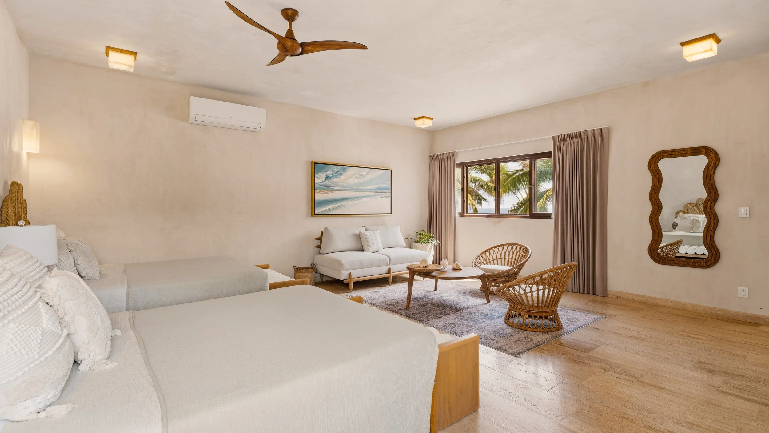 A bright and airy hotel room with two beds, a seating area with a sofa and wicker chairs, a large window with curtains showing palm trees outside, and tropical decor including a beach-themed painting and a mirror.