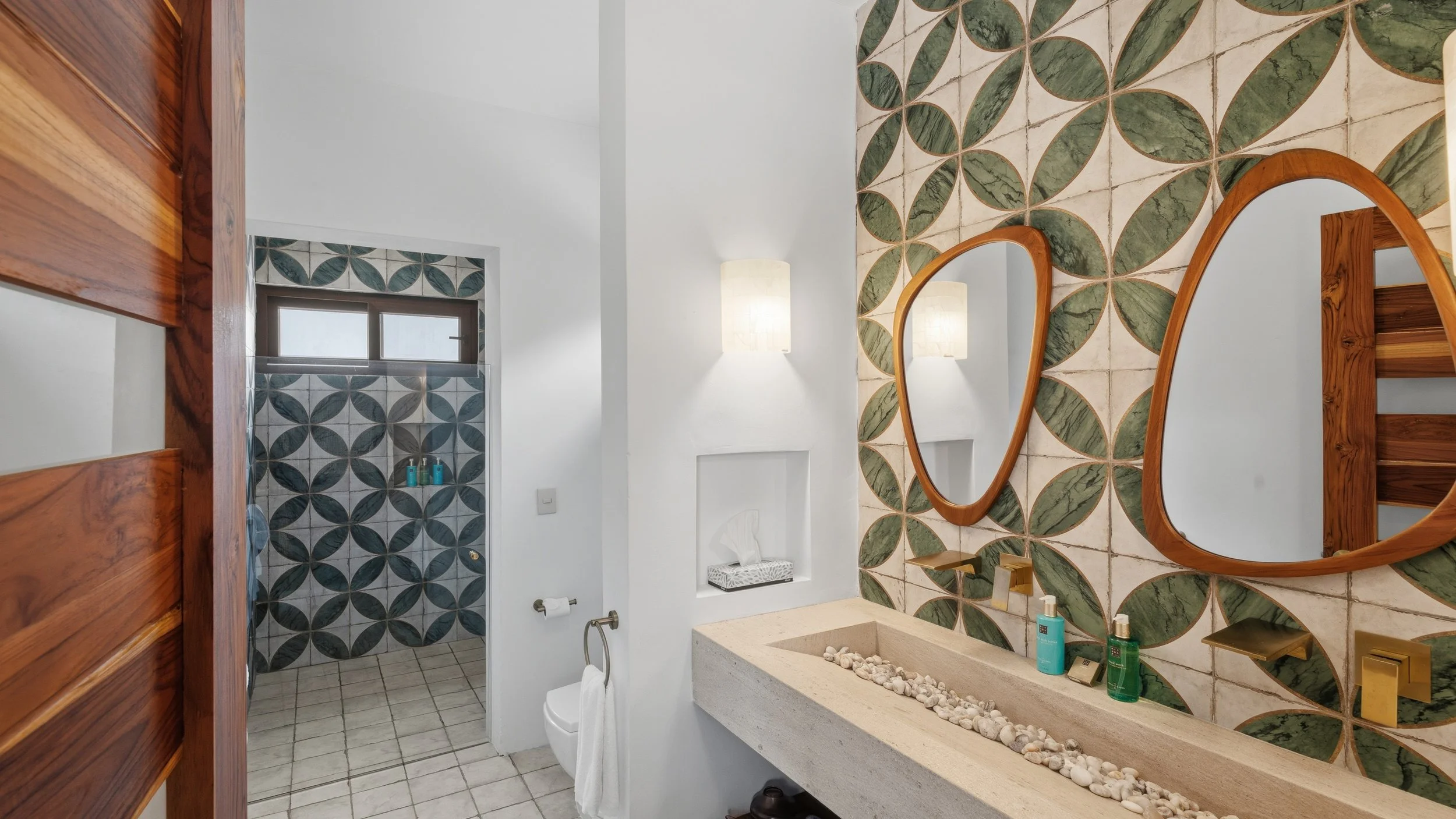 Bathroom with patterned tile wall, wooden framed mirror, stone sink with pebbles, and shower area with geometric tile floor and a small window.