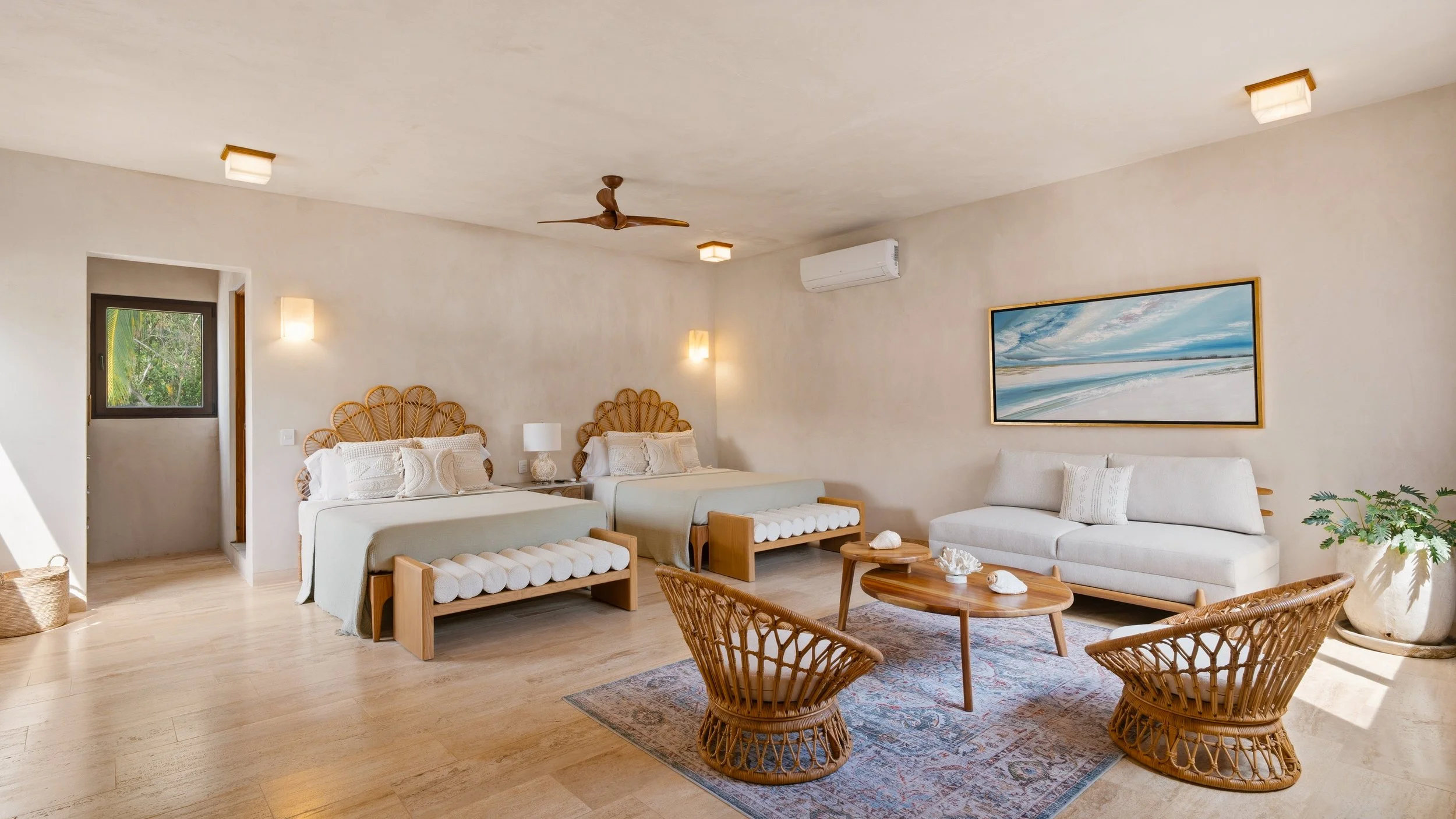 Interior of a living room with white walls, wooden furniture, wicker chairs, framed wall art of a beach scene, and a ceiling fan.