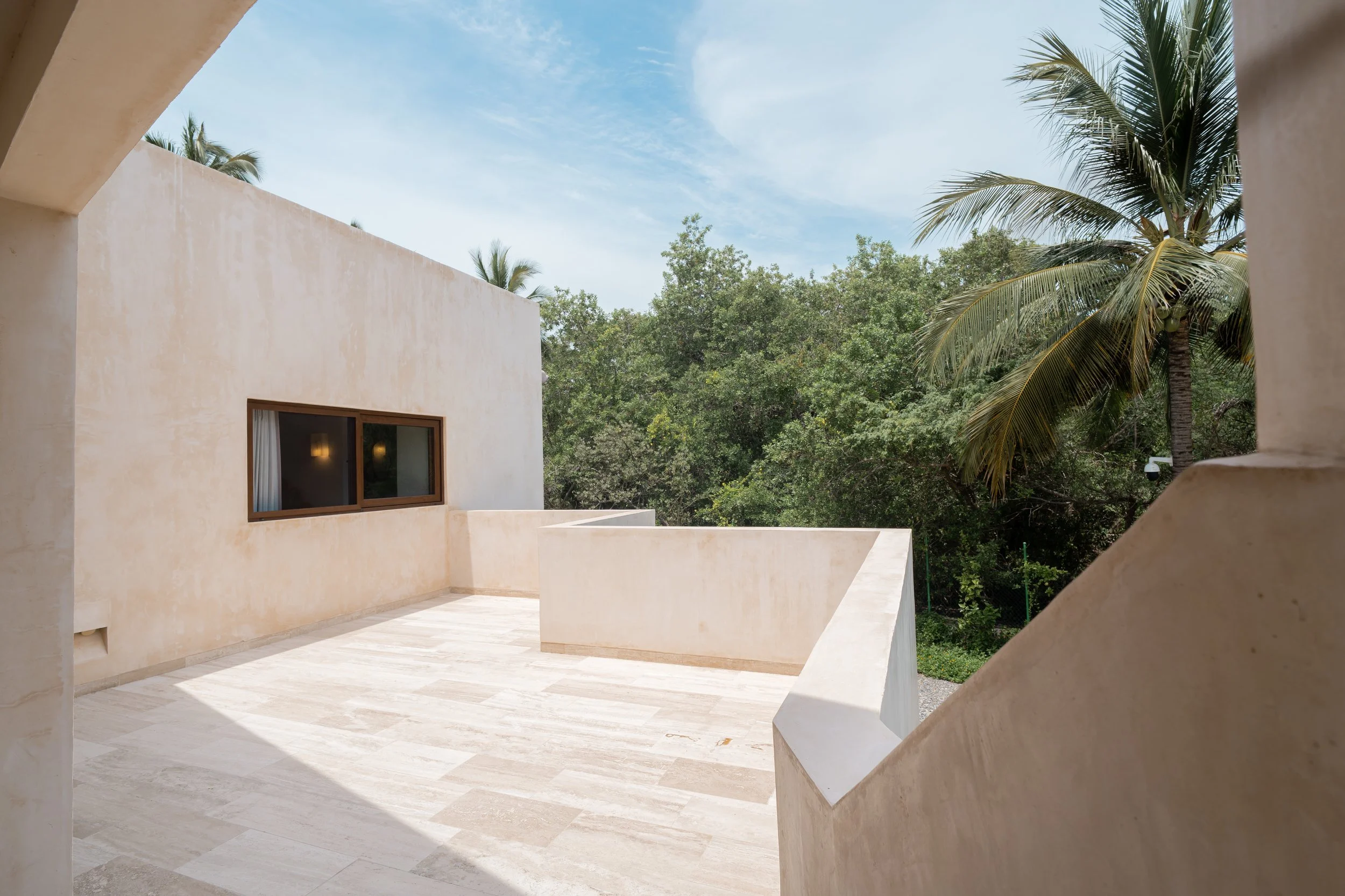 View from a balcony with beige walls, a window, and trees including palm trees under a blue sky.