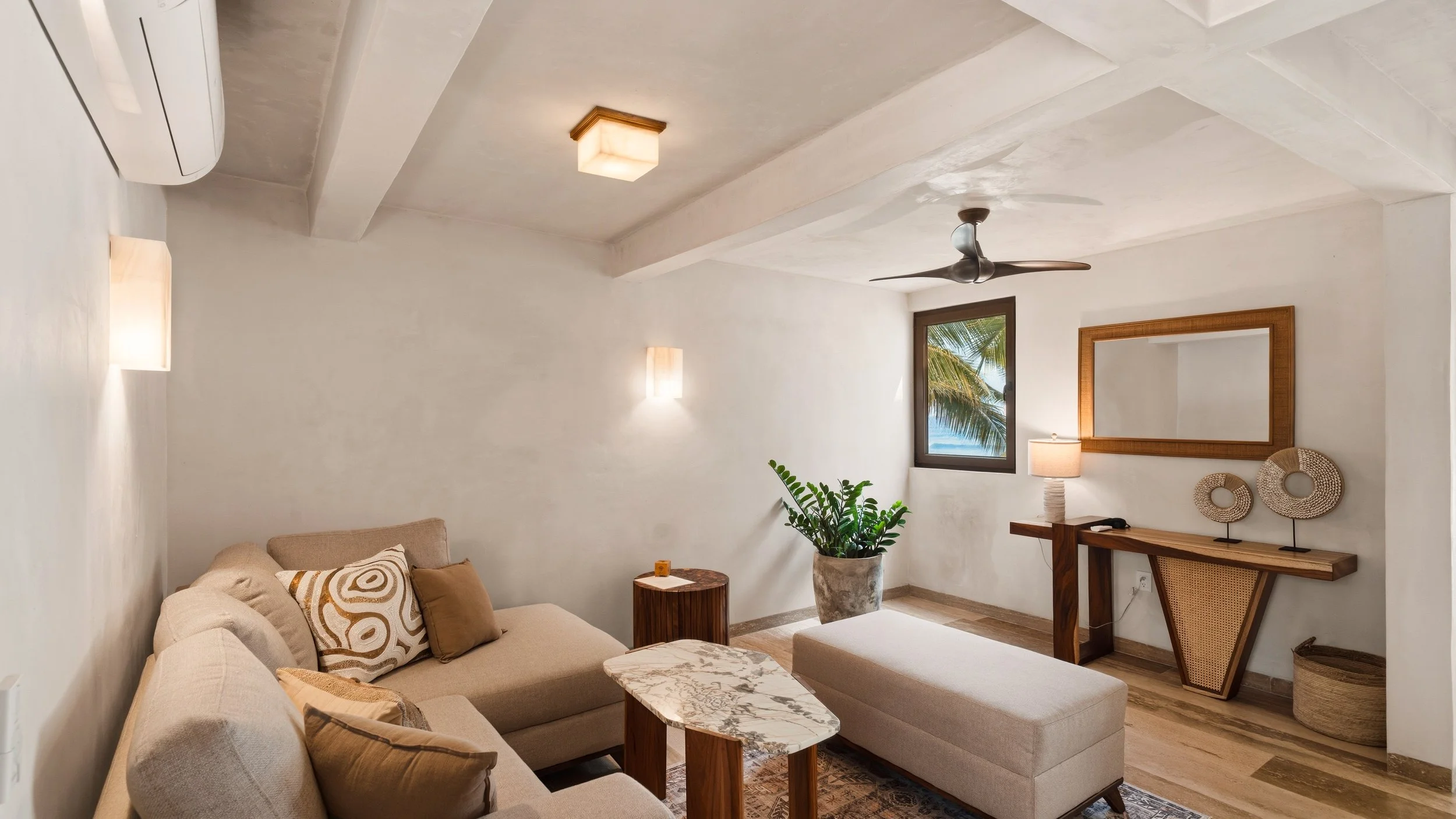 Living room with beige sectional sofa, patterned throw pillows, wooden side table, large potted plant, wall-mounted mirror, table lamp, window with palm tree view, ceiling fan, and modern ceiling lights.