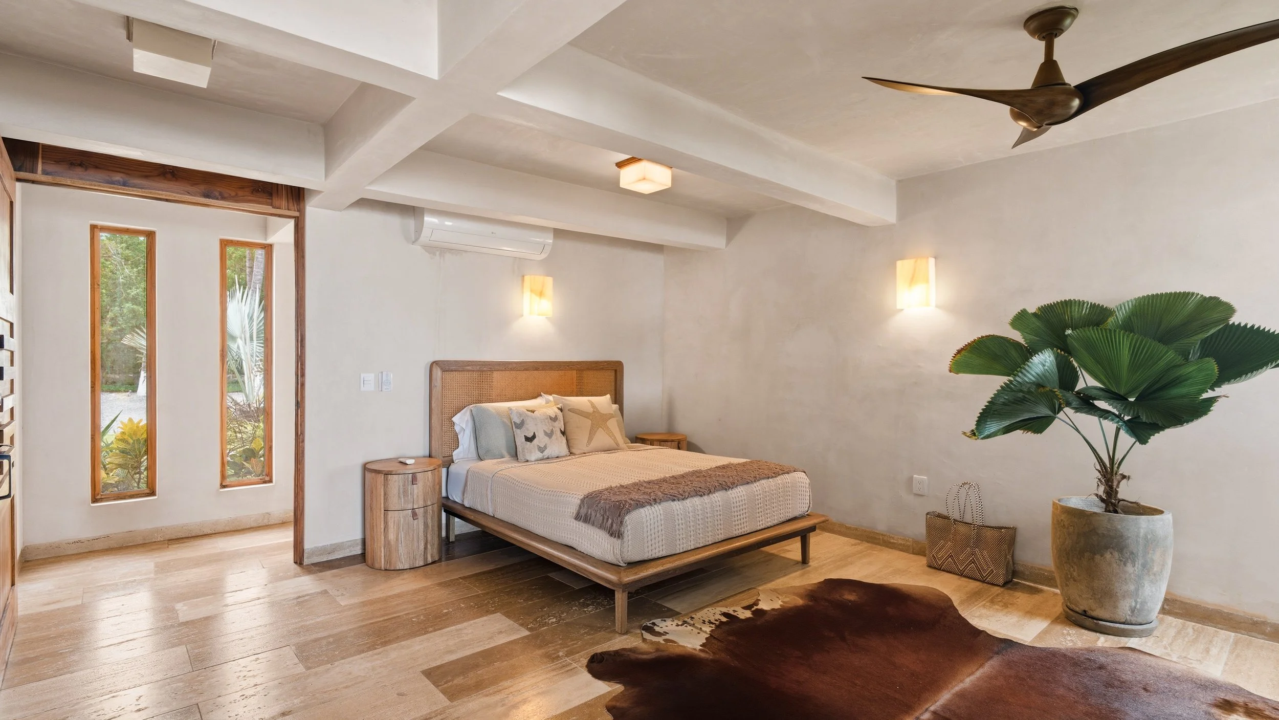 A bedroom with a wooden bed frame, beige bedding, and decorative pillows featuring starfish and bird designs. There are two small wooden nightstands, an air conditioning unit, and a large leafy plant in a pot. The room has white textured walls, warm 
