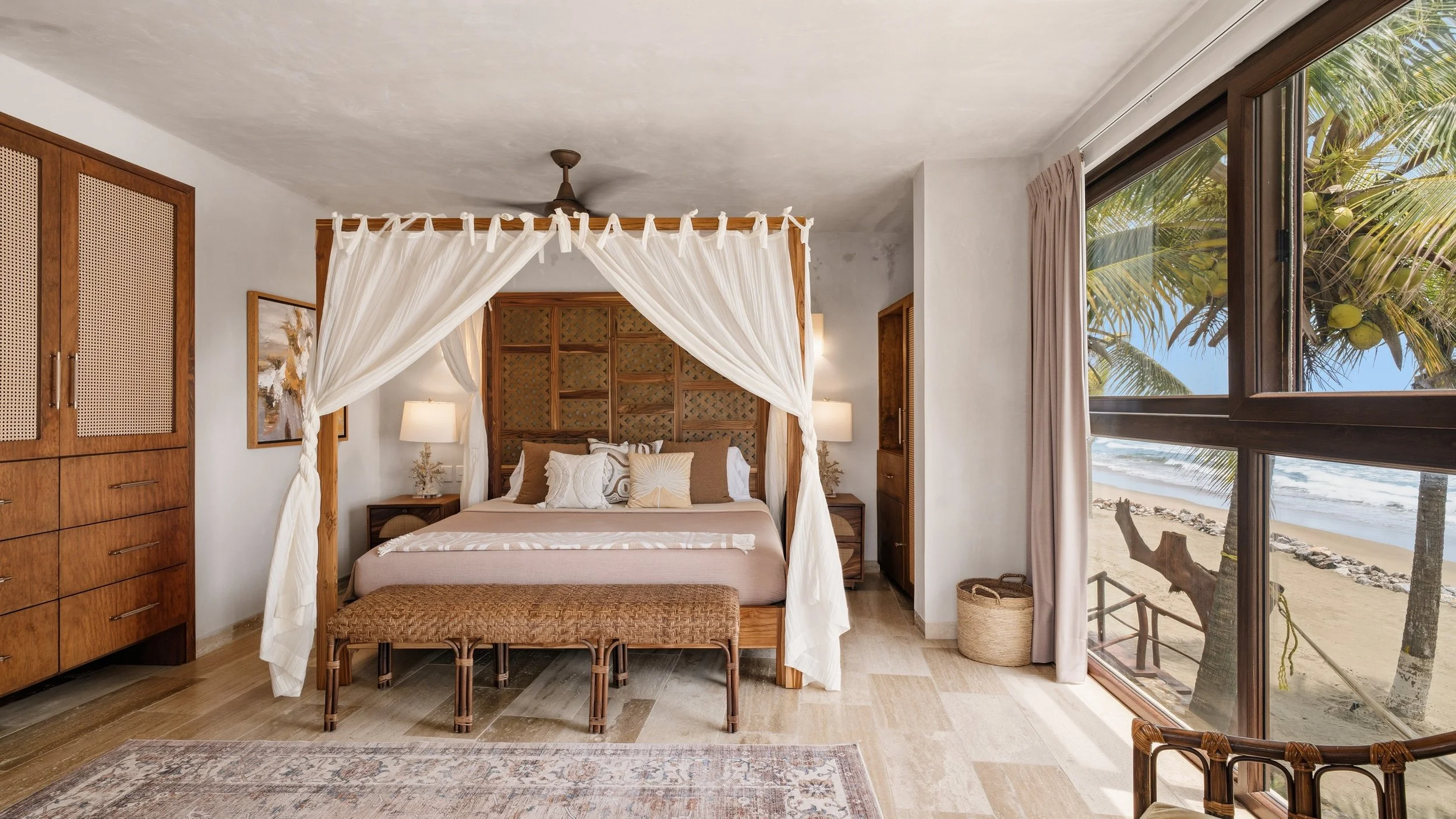 A bedroom with a four-poster bed and white curtains, wooden furniture, and a large window showing a beach and palm trees outside.
