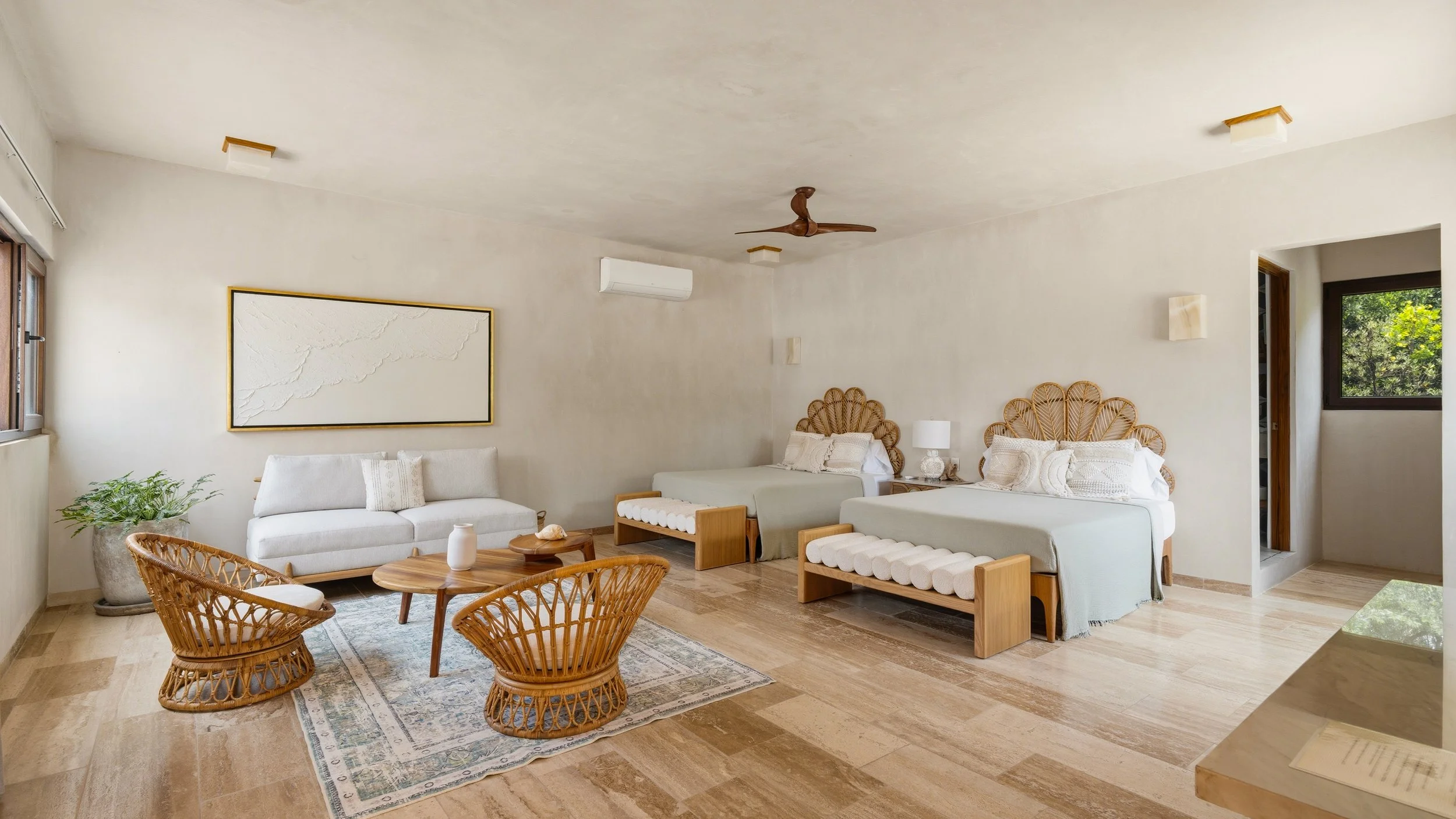 A bright and airy living room with two beds, a white sofa, rattan chairs, a wooden coffee table, and large windows showing greenery outside.