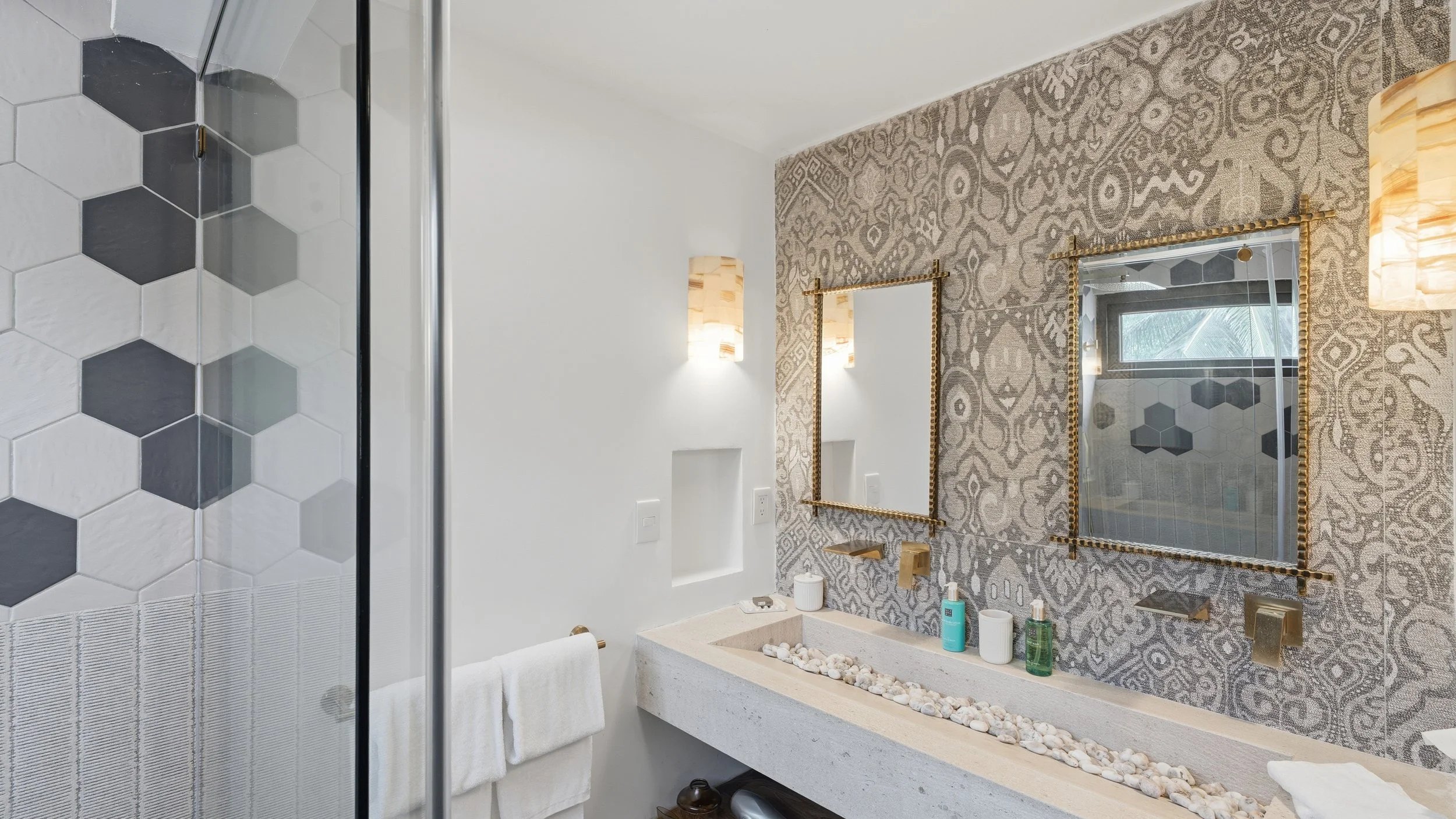 Bathroom with patterned wallpaper, two gold-framed mirrors, a sink with toiletries, a towel, and a decorative rocky basin. Part of the shower with hexagonal tiles is visible.
