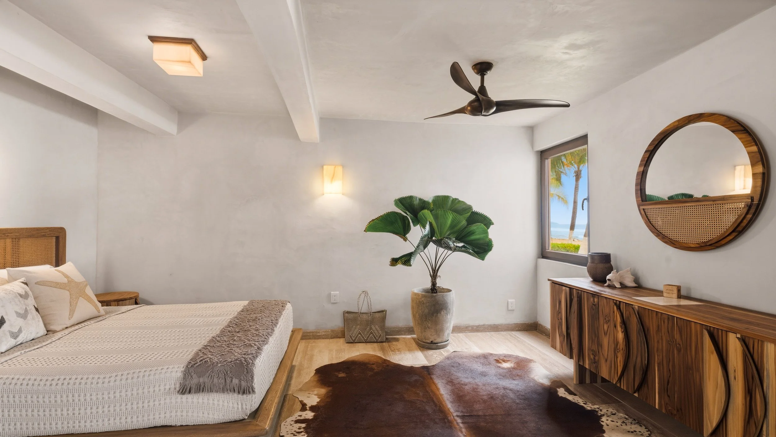 A bedroom with white walls, a wooden bed with starfish pillows, a large potted plant, a cowhide rug, a wooden sideboard with decor, a round mirror, a window showing palm trees and the beach, ceiling fan, and wall-mounted light.