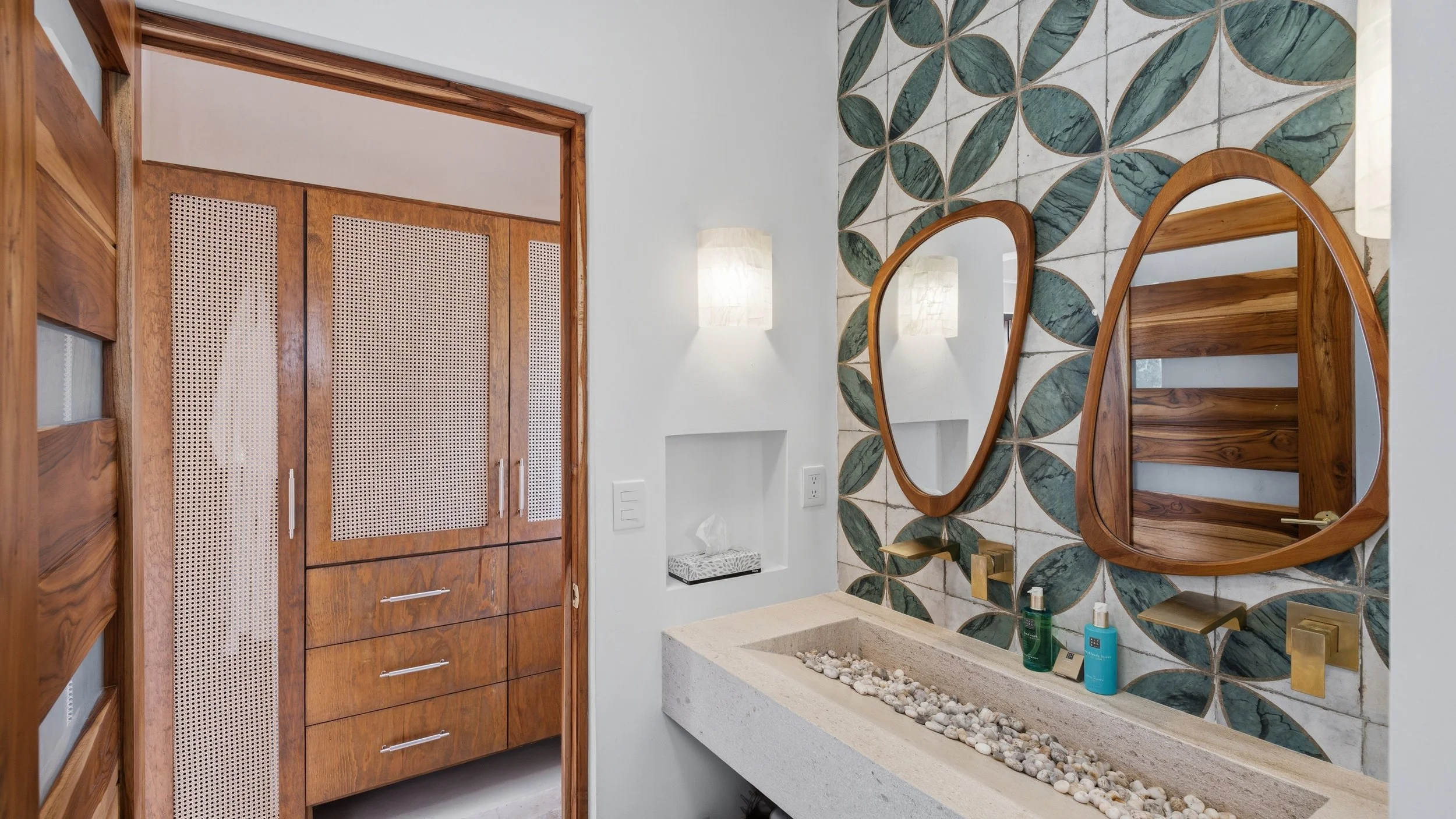 Bathroom with a large mirror, green leaf-patterned tile wall, wooden storage cabinets, and a concrete sink with pebbles inside.