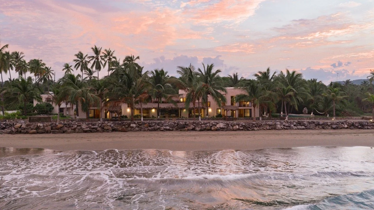 A tropical beachfront resort at sunset with palm trees, a stone wall, and a building with lit windows, ocean waves in the foreground, and a colorful sky.