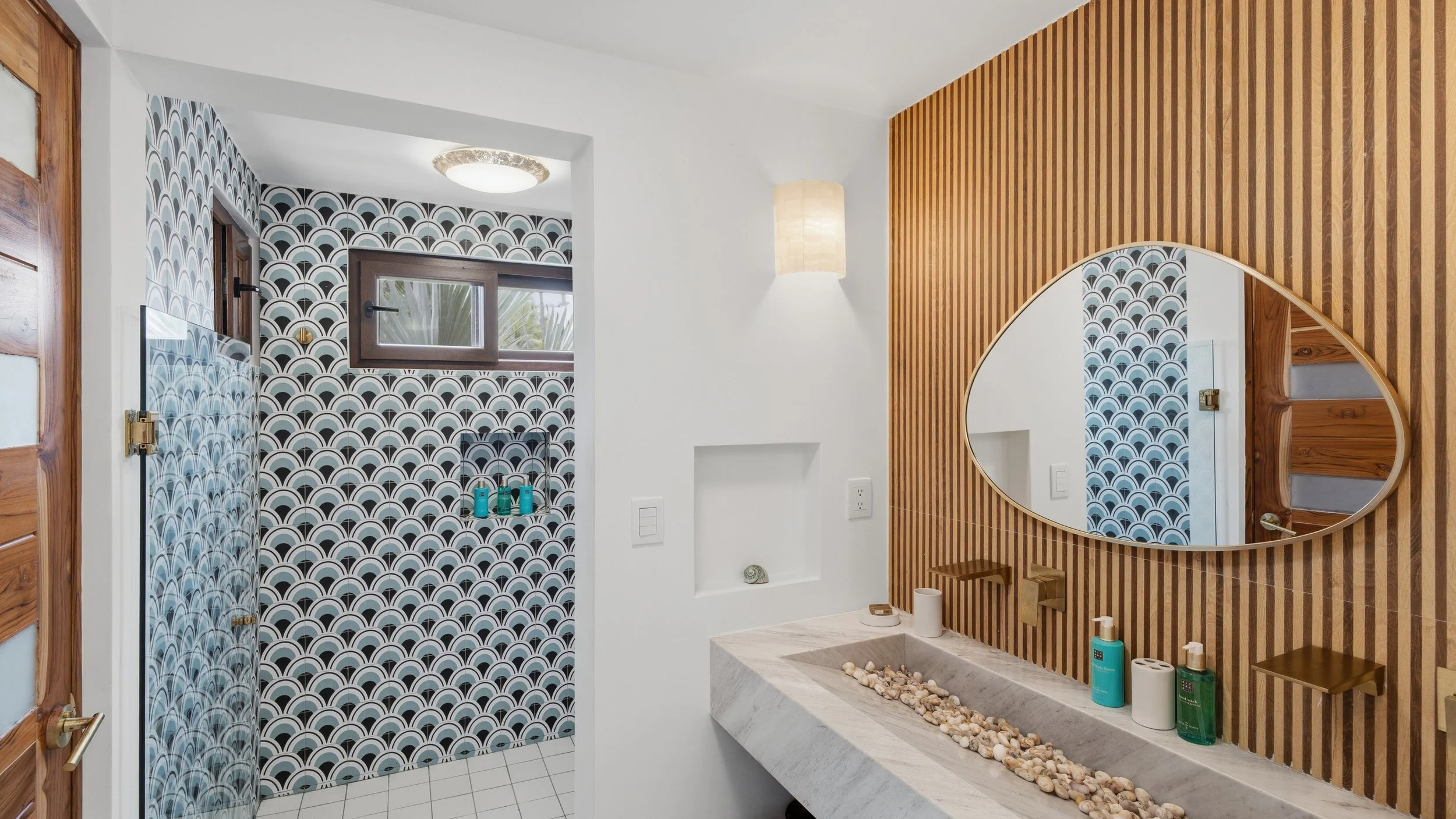 Modern bathroom with a patterned shower area, wooden accents, and a large oval mirror above a stone sink filled with rocks.
