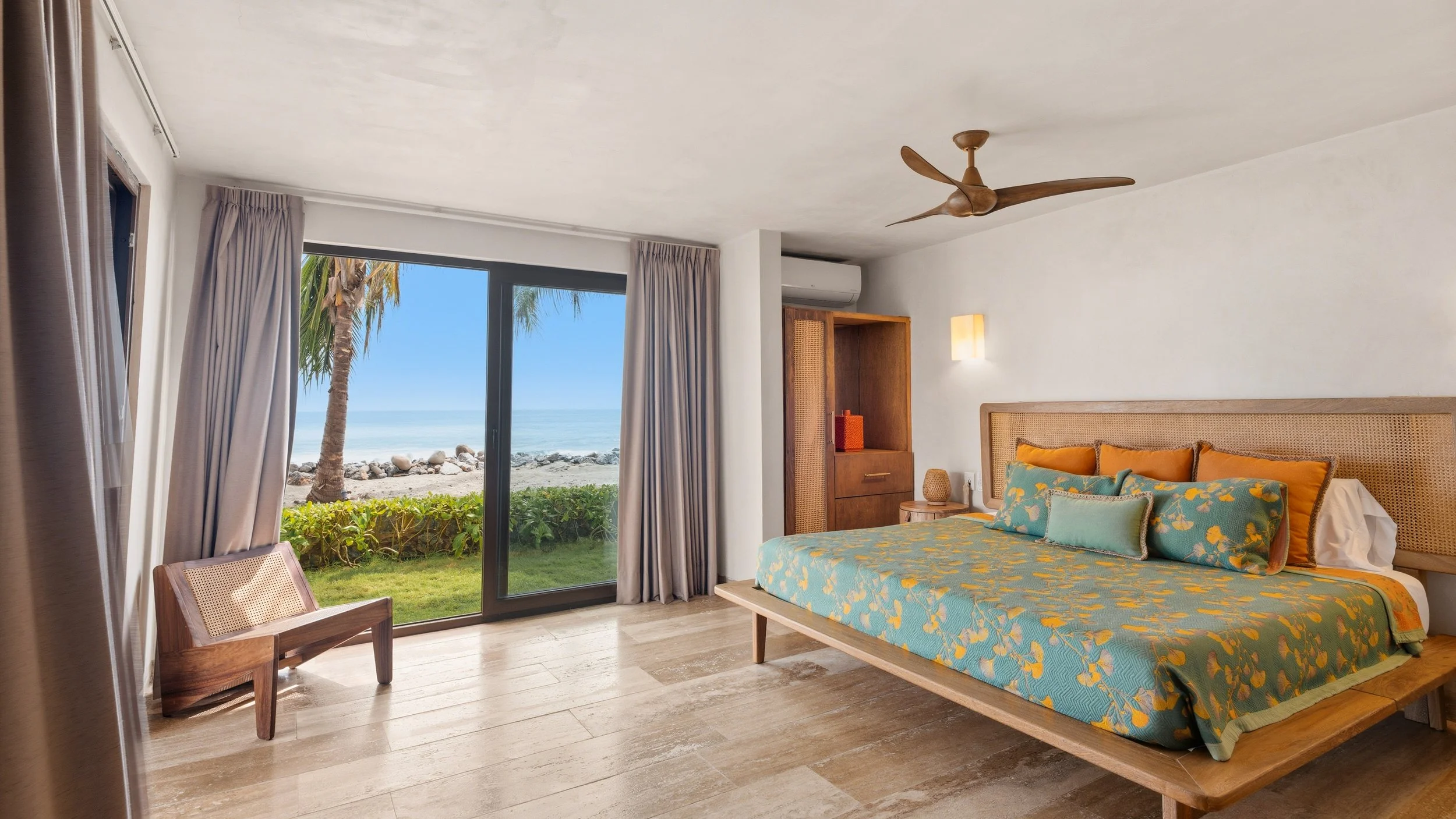 A bedroom with a sliding glass door leading to a beach view. The room has a bed with floral bedding, a wooden headboard, and a small wooden chair near the window. Outside, palm trees and rocks line the beach.