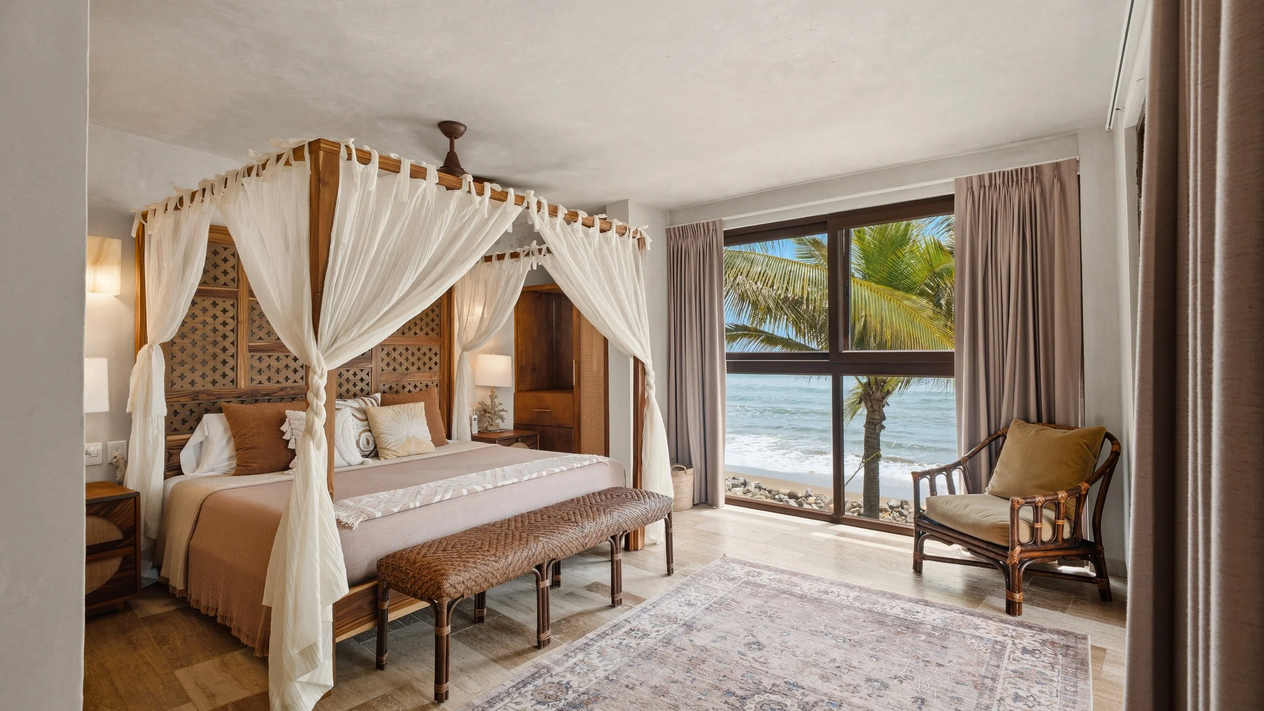 A bedroom with a large four-poster bed with white curtains. There are beach views outside the window, with palm trees and ocean visible.
