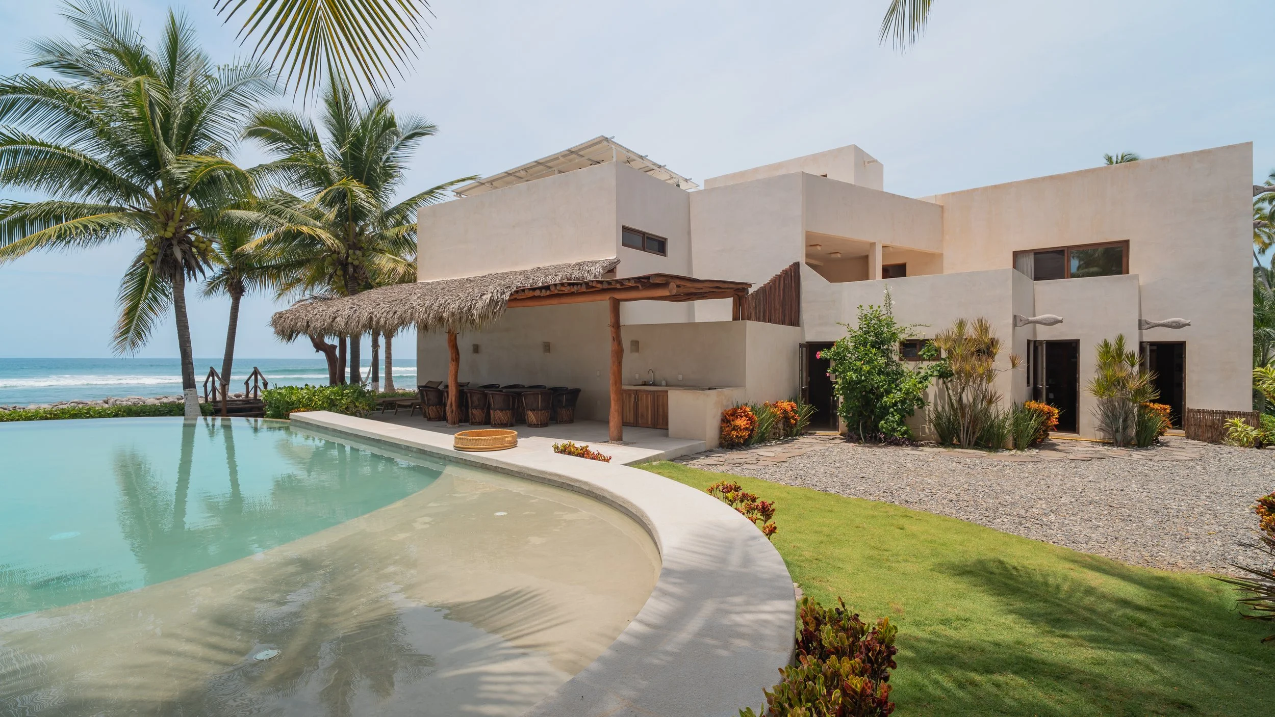 Modern beachfront house with outdoor kitchen, tiki hut, swimming pool, palm trees, and ocean in the background.