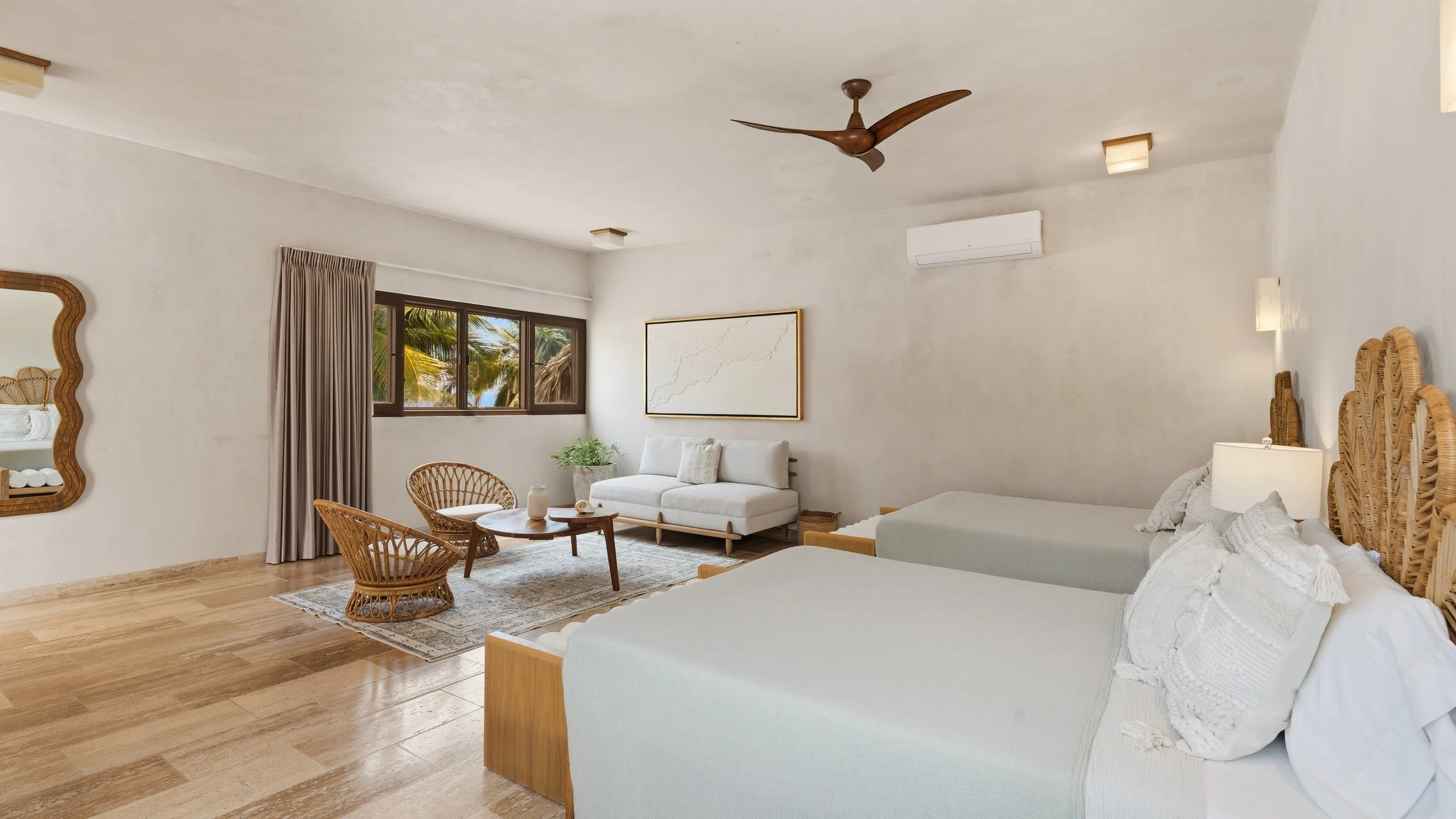 Bright living room with white walls, wooden furniture, rattan chairs, a couch, and a bed. Large window with curtains shows palm trees outside. Ceiling fan and air conditioner visible.