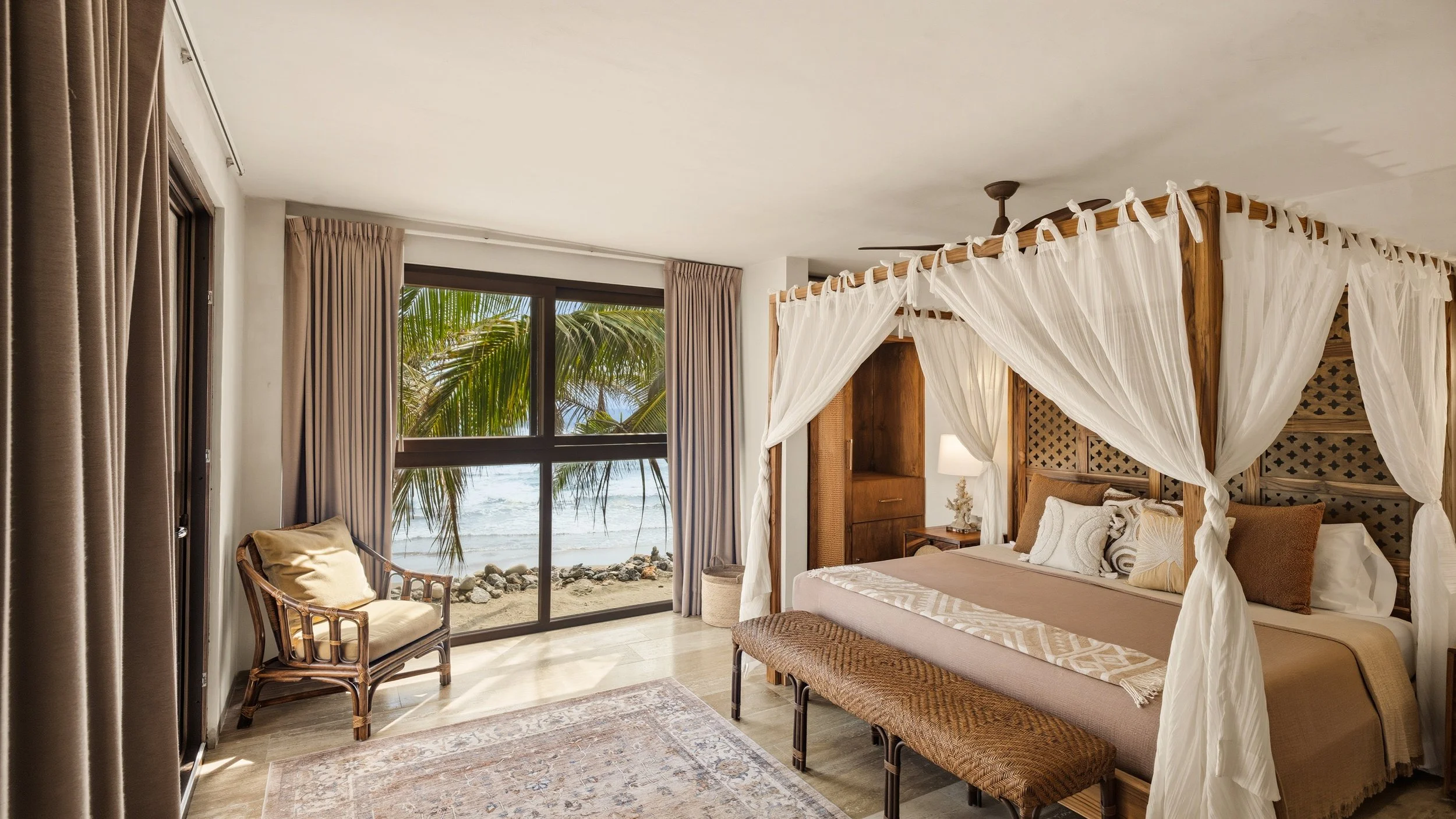 Interior of a beachside bedroom with a four-poster bed draped in white curtains, positioned near large glass sliding doors showing palm trees and the ocean outside, with beige curtains framing the doors and a wicker chair with a cushion nearby.