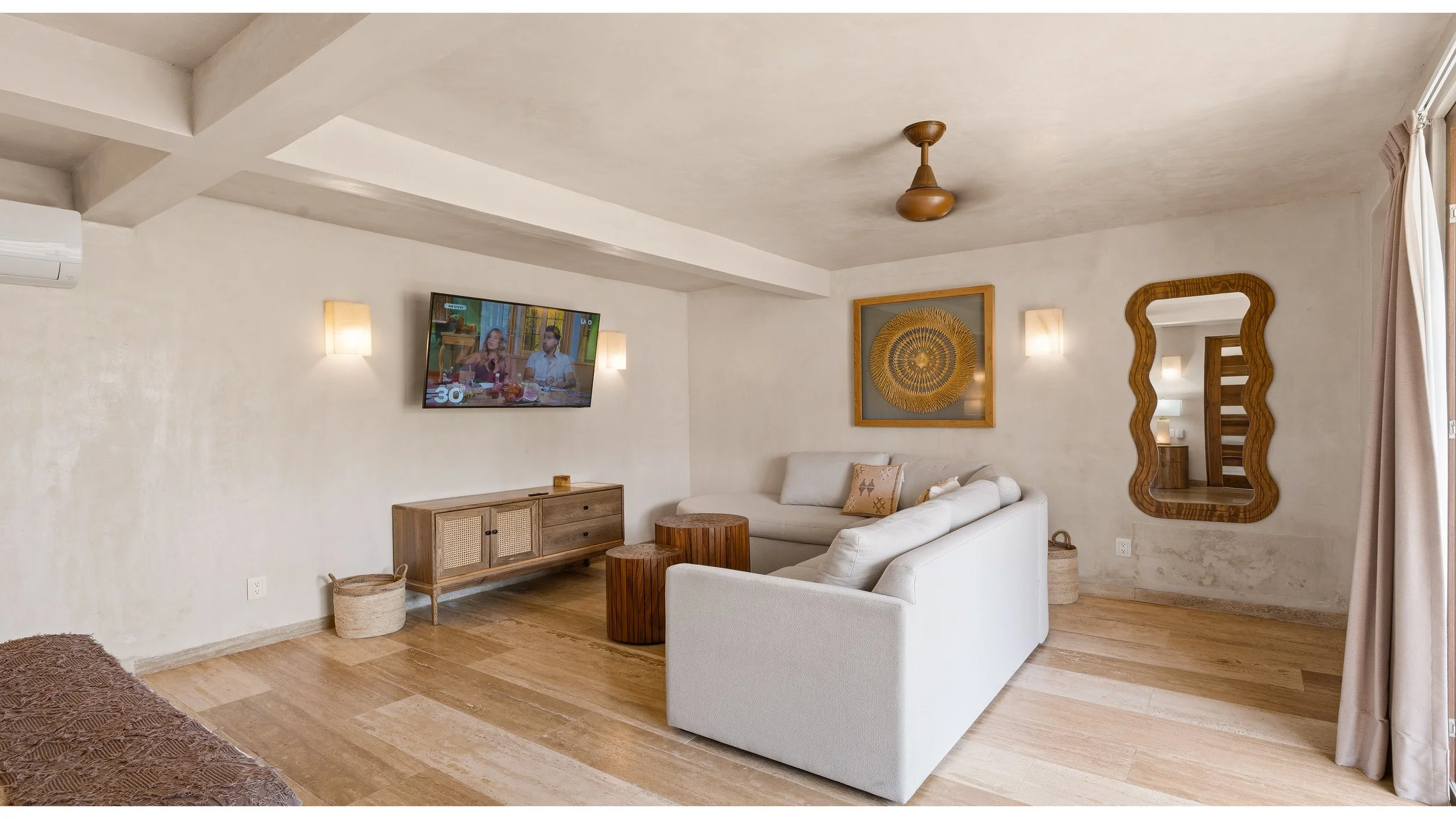 Living room with cream-colored sofas, wooden side tables, a wooden TV stand with a TV mounted on the wall, decorative wall art, a vertical mirror with a wavy wooden frame, and curtains on the right window.
