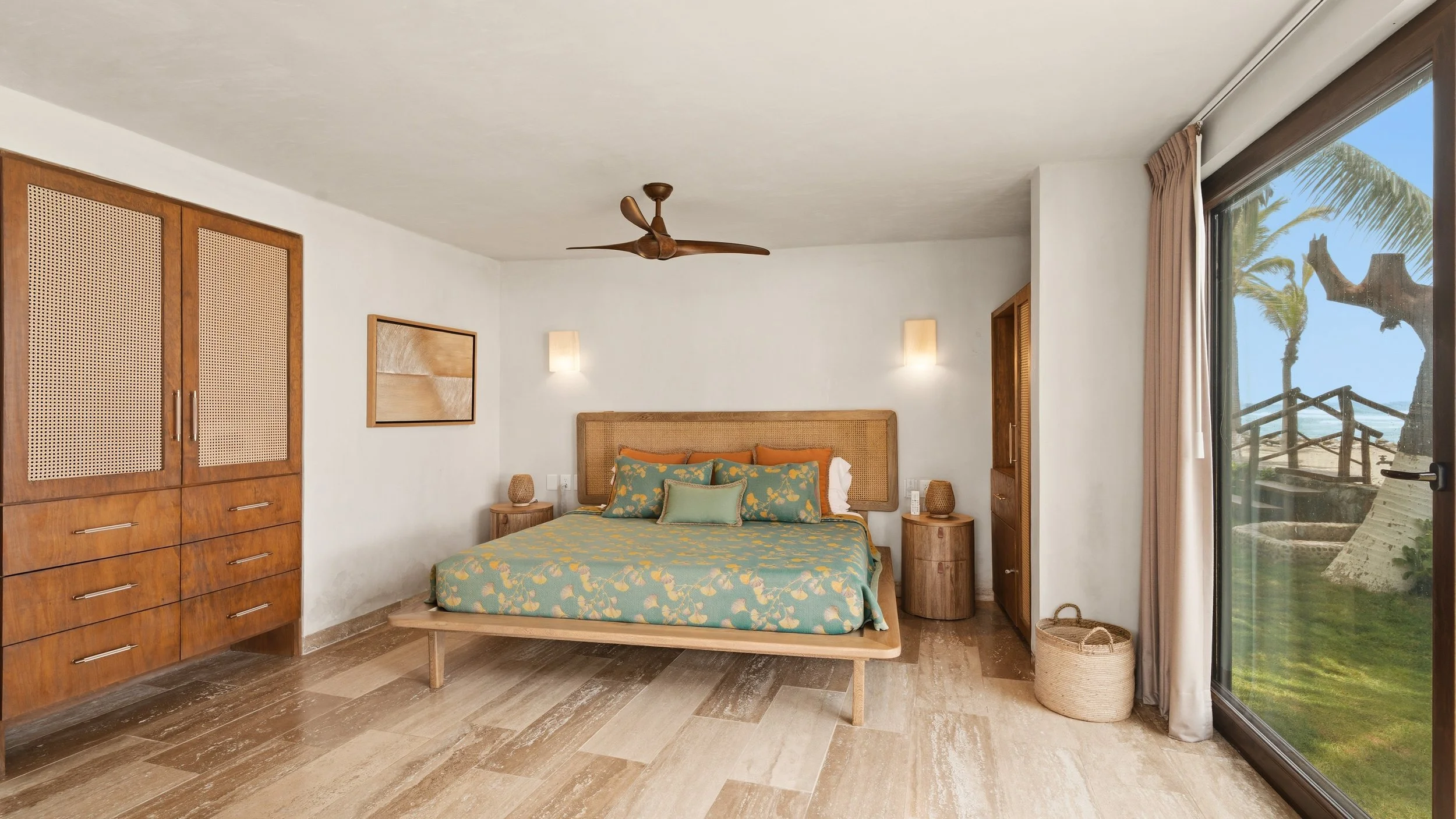 A bedroom with a wooden bed, patterned bedding, and two nightstands. To the right, there is a large glass door with a view of palm trees and a beach outside.