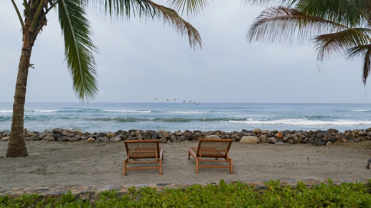 Two wooden lounge chairs on a sandy beach facing the ocean, with palm trees on either side and rocks along the shoreline, under a cloudy sky.