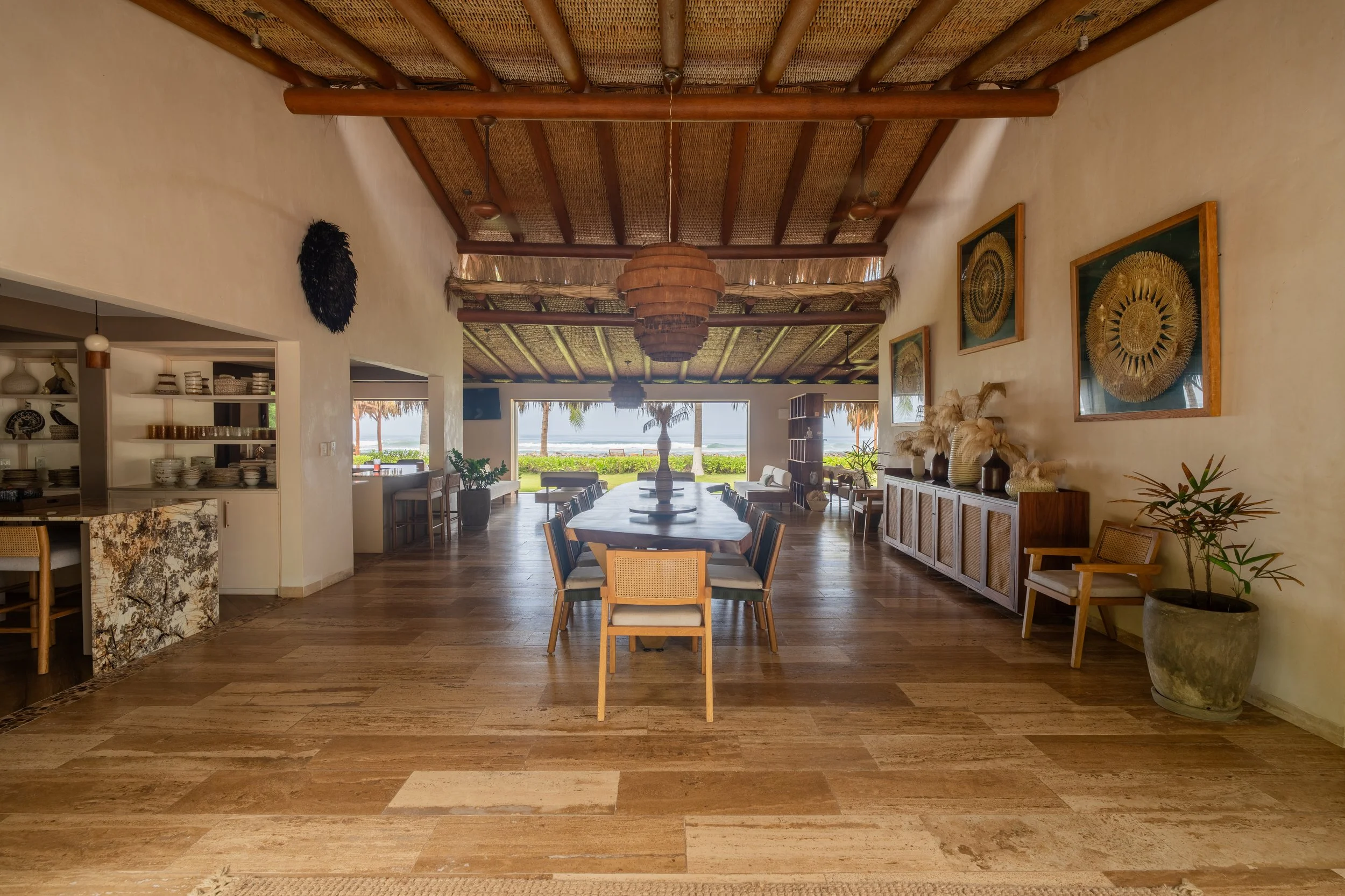 Interior of a beachside restaurant with wooden furniture, decorative wall art, and an open view of palm trees and the ocean in the background.