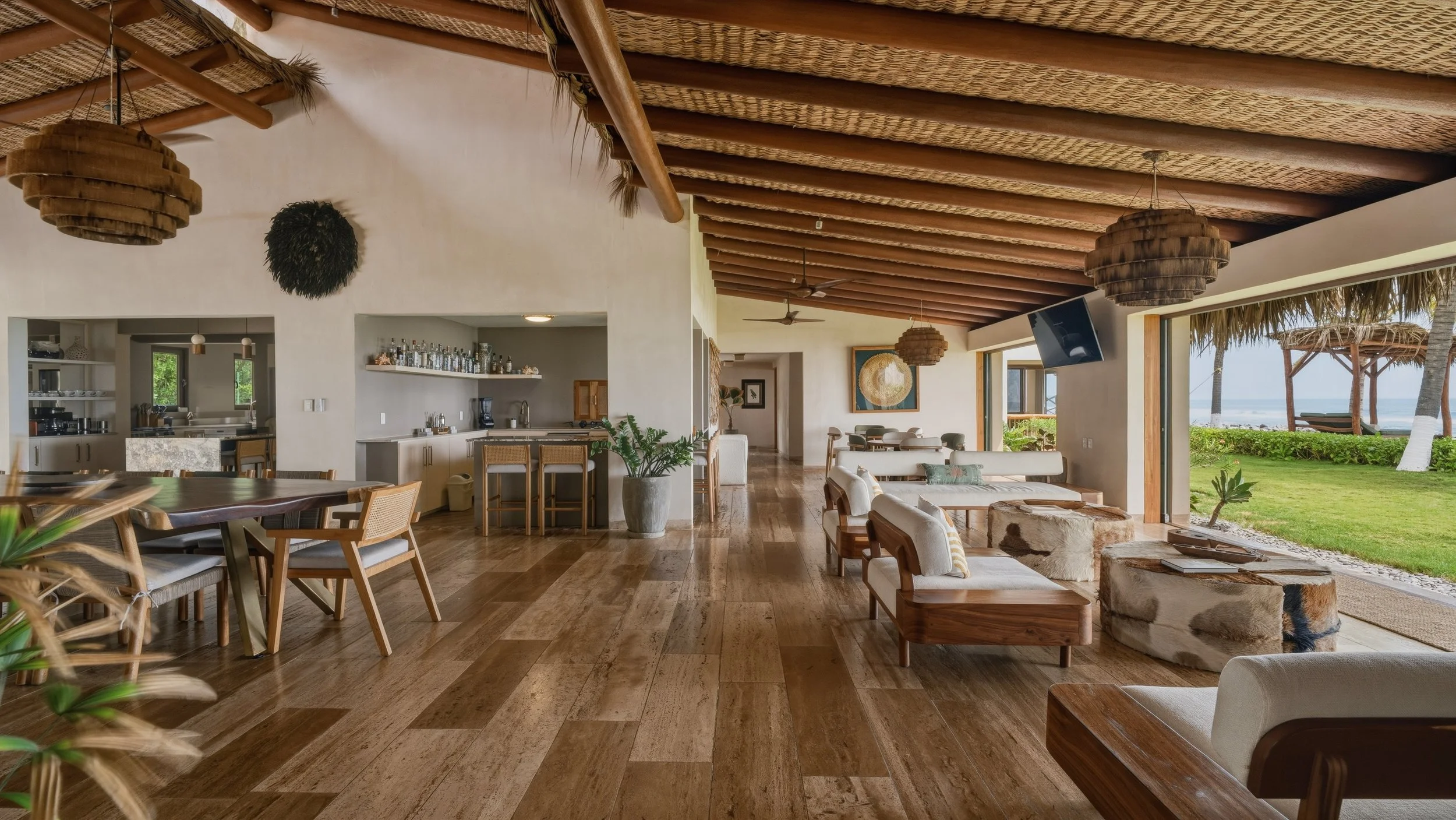 Interior of a tropical beach house with wood flooring, white walls, and open sliding doors leading to a grassy area with palm trees and a beach in the background. Contains a dining table, bar area, and seating with ocean views.