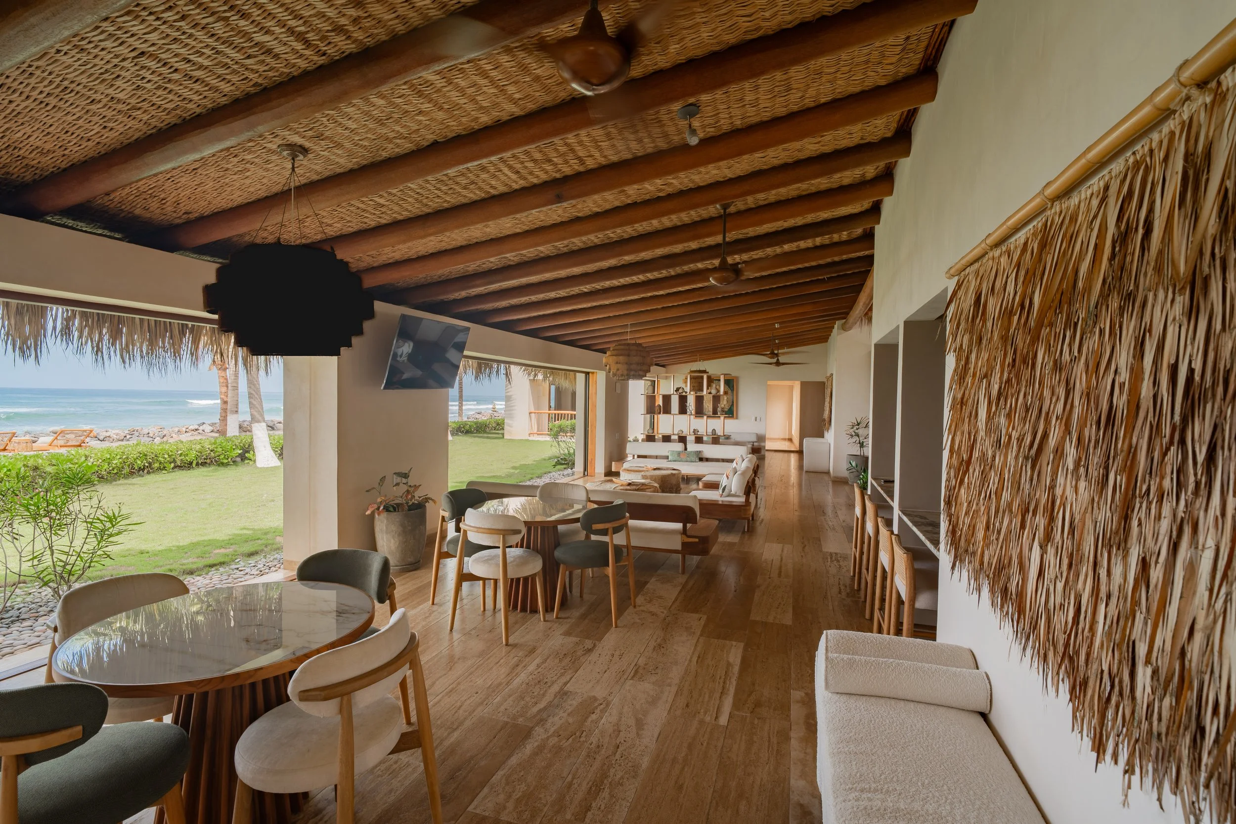 Interior of a beachside restaurant or lounge with a thatched roof, wooden beams, and large open windows with a view of the beach and ocean.