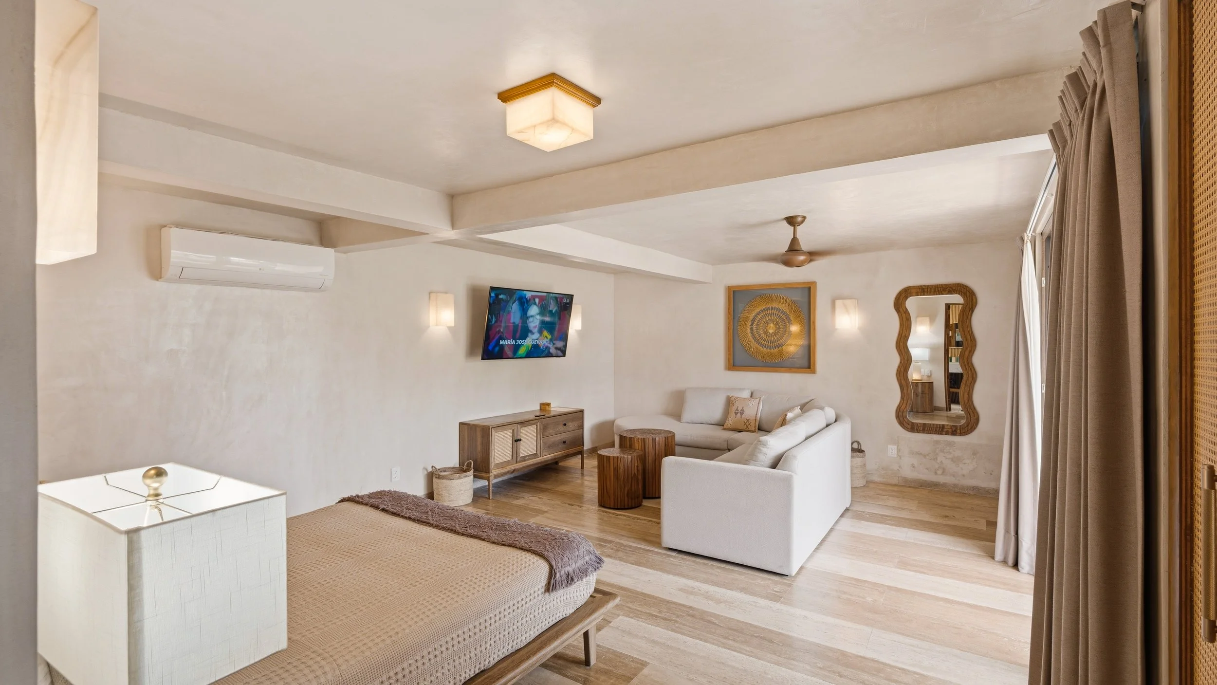 A living room with beige walls, a white sectional sofa, a wooden TV console with a TV, a wall mirror, and a window with beige curtains.