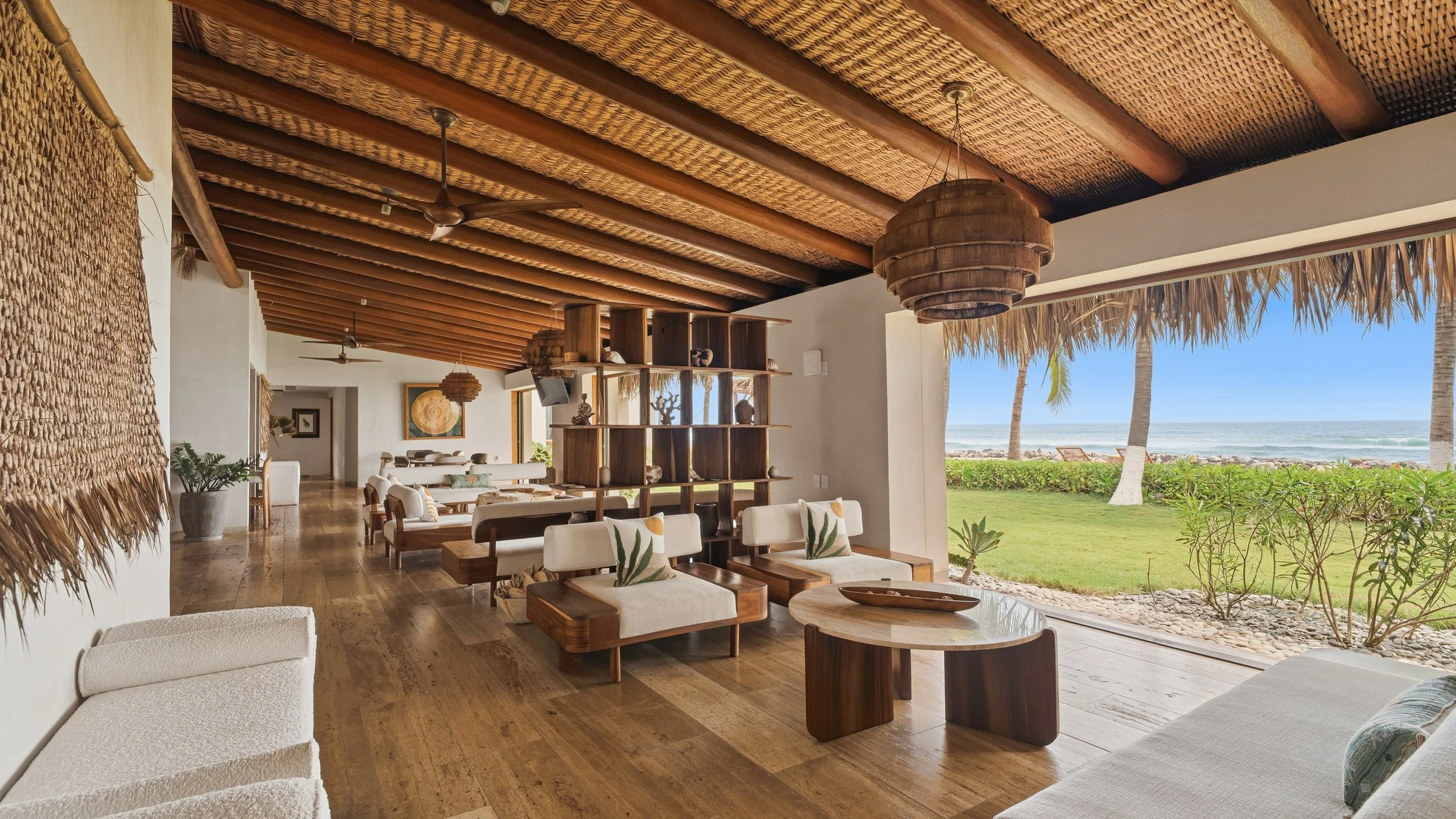 Open-air lounge area with tropical decor overlooking the beach, featuring wooden furniture, woven ceiling fans, and a view of the ocean and palm trees.