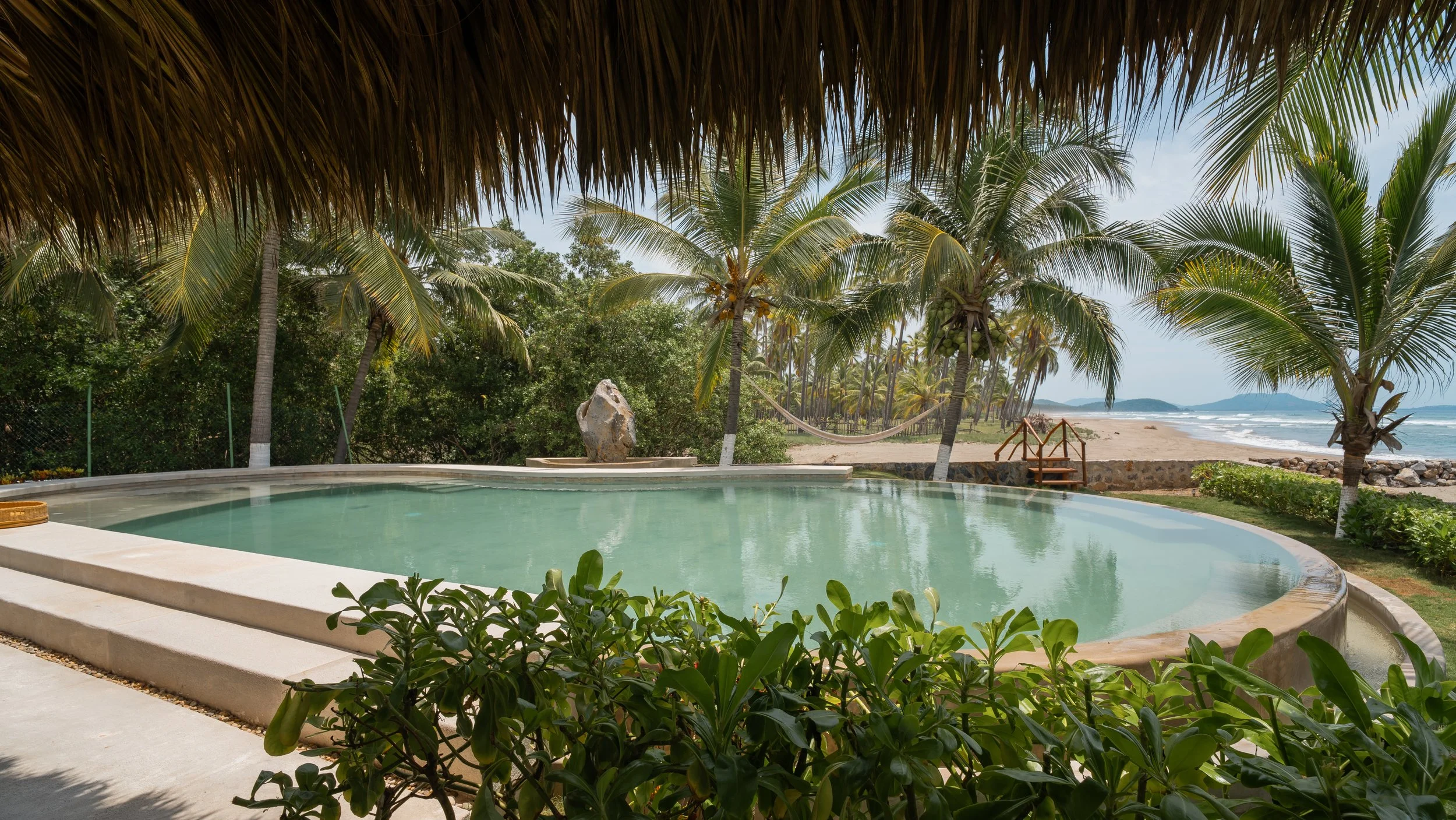 A swimming pool with a view of palm trees and a sandy beach by the ocean, under a thatched roof.
