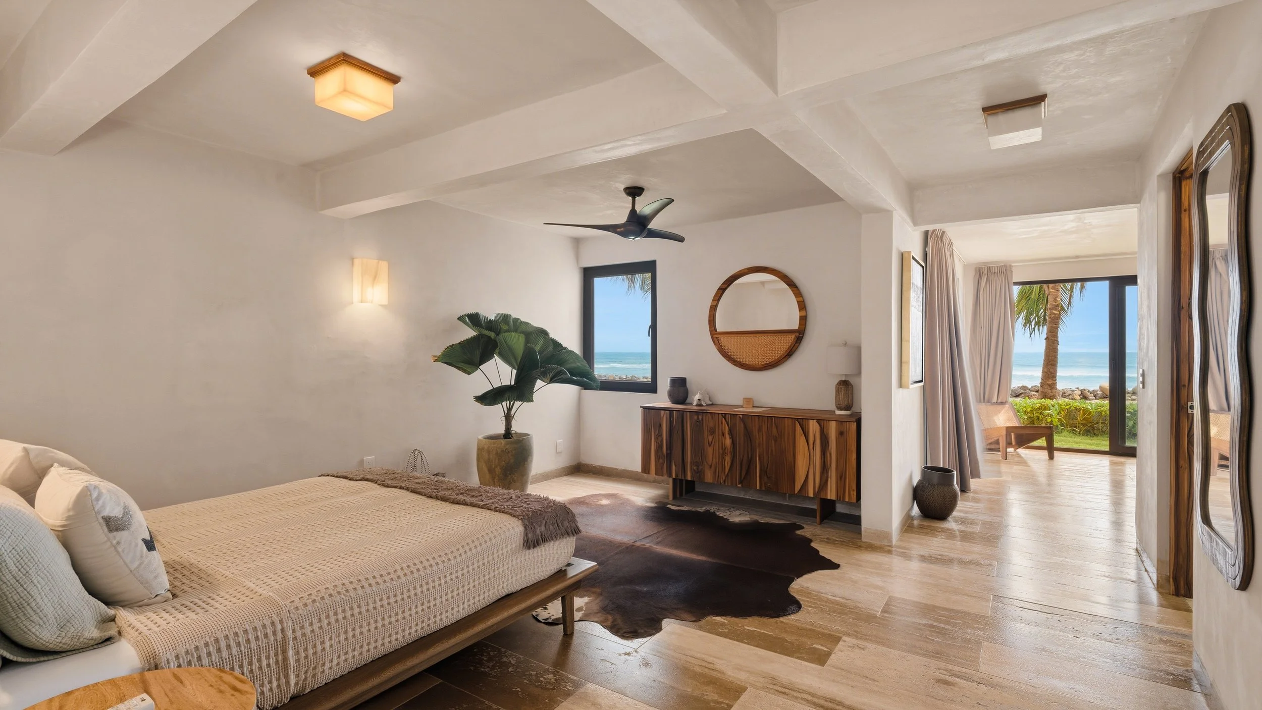 A bedroom with a large bed, wooden dresser, potted plant, and windows showing a view of the ocean and palm trees outside.