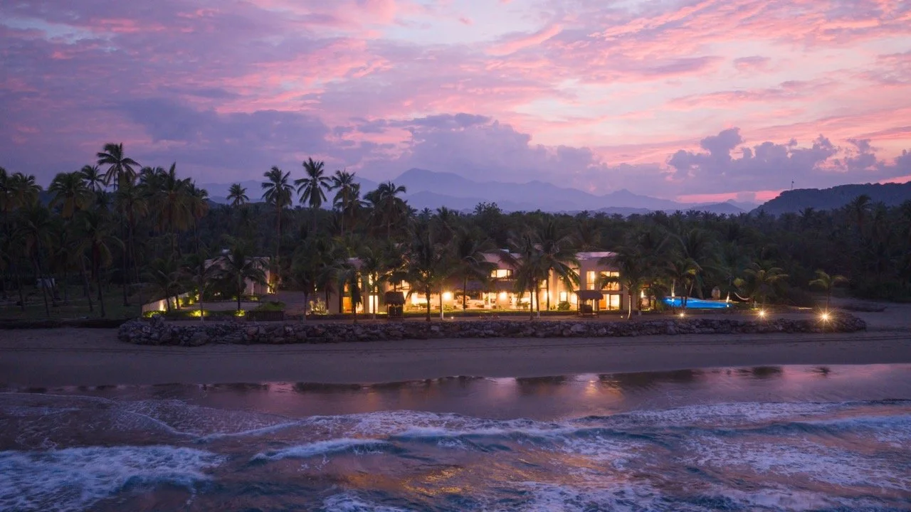 A tropical beachside resort at dusk with palm trees, a sandy beach, and ocean waves.