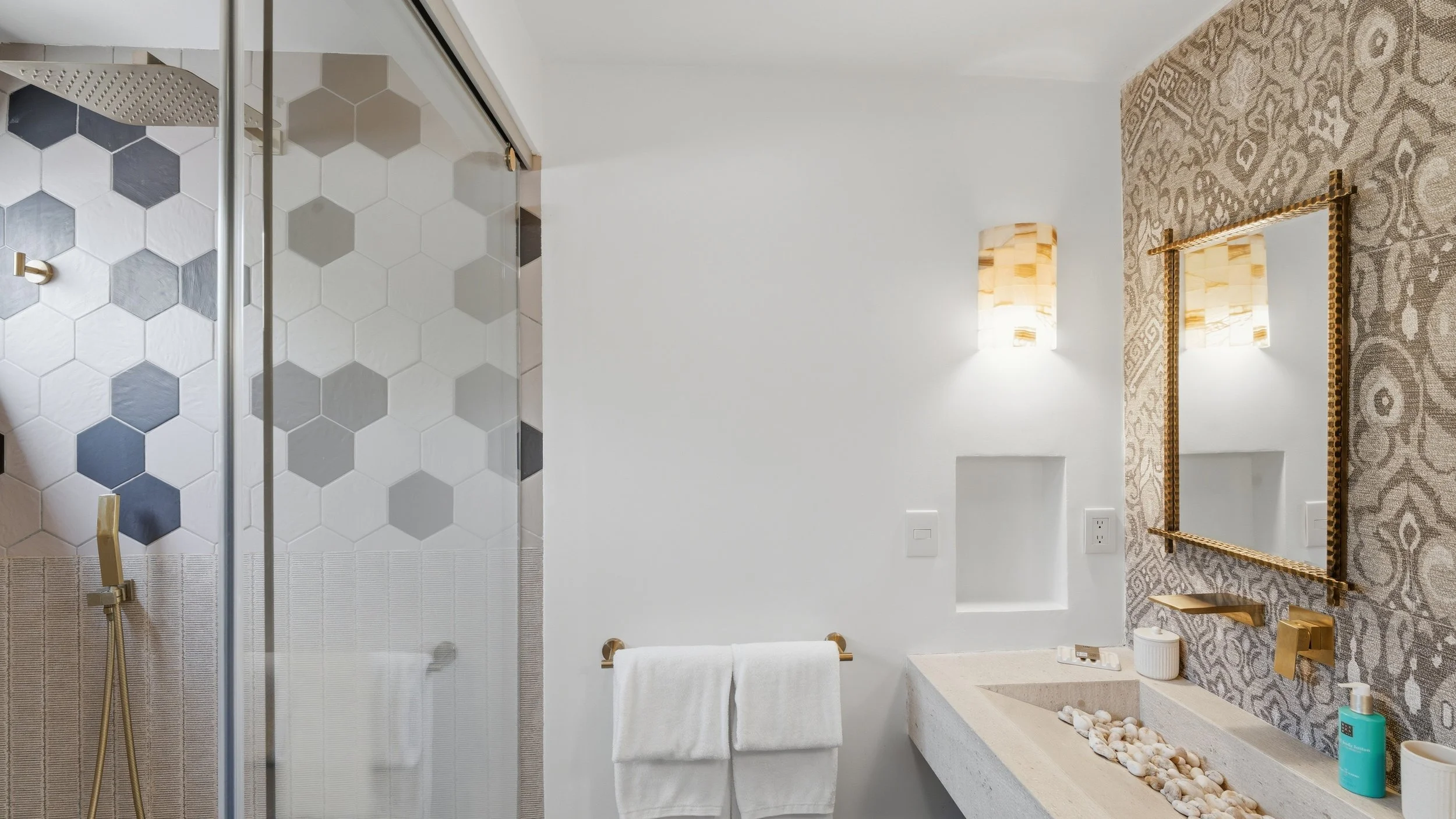 Modern bathroom with shower area featuring hexagonal tiles in shades of white, gray, and dark gray. The vanity has a large mirror with a gold frame, two wall-mounted light fixtures, and a patterned wallpaper behind the mirror. There is a concrete cou