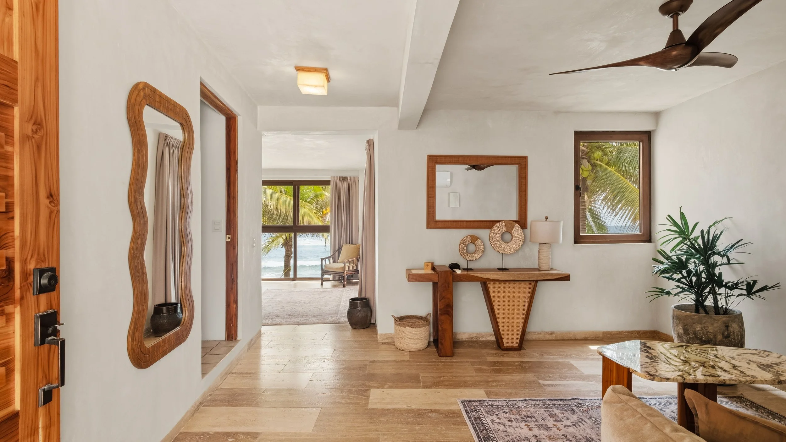 Interior view of a room with white walls, wooden accents, a mirror, plants, and a view outside of palm trees and the ocean through windows.