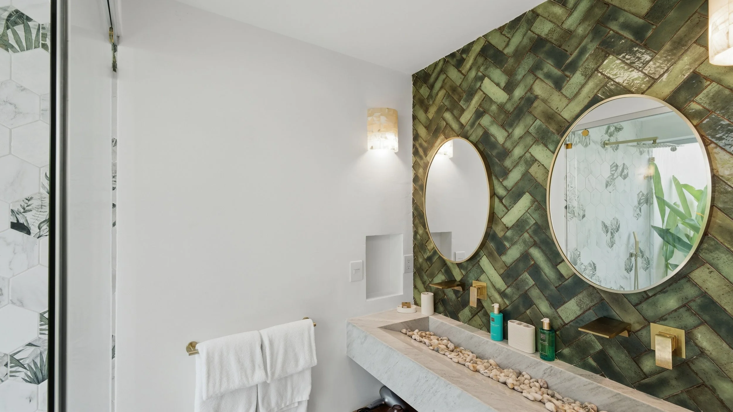 Modern bathroom with green herringbone tiled wall, oval mirrors, and decorative sink with rocks, candles, and toiletries.