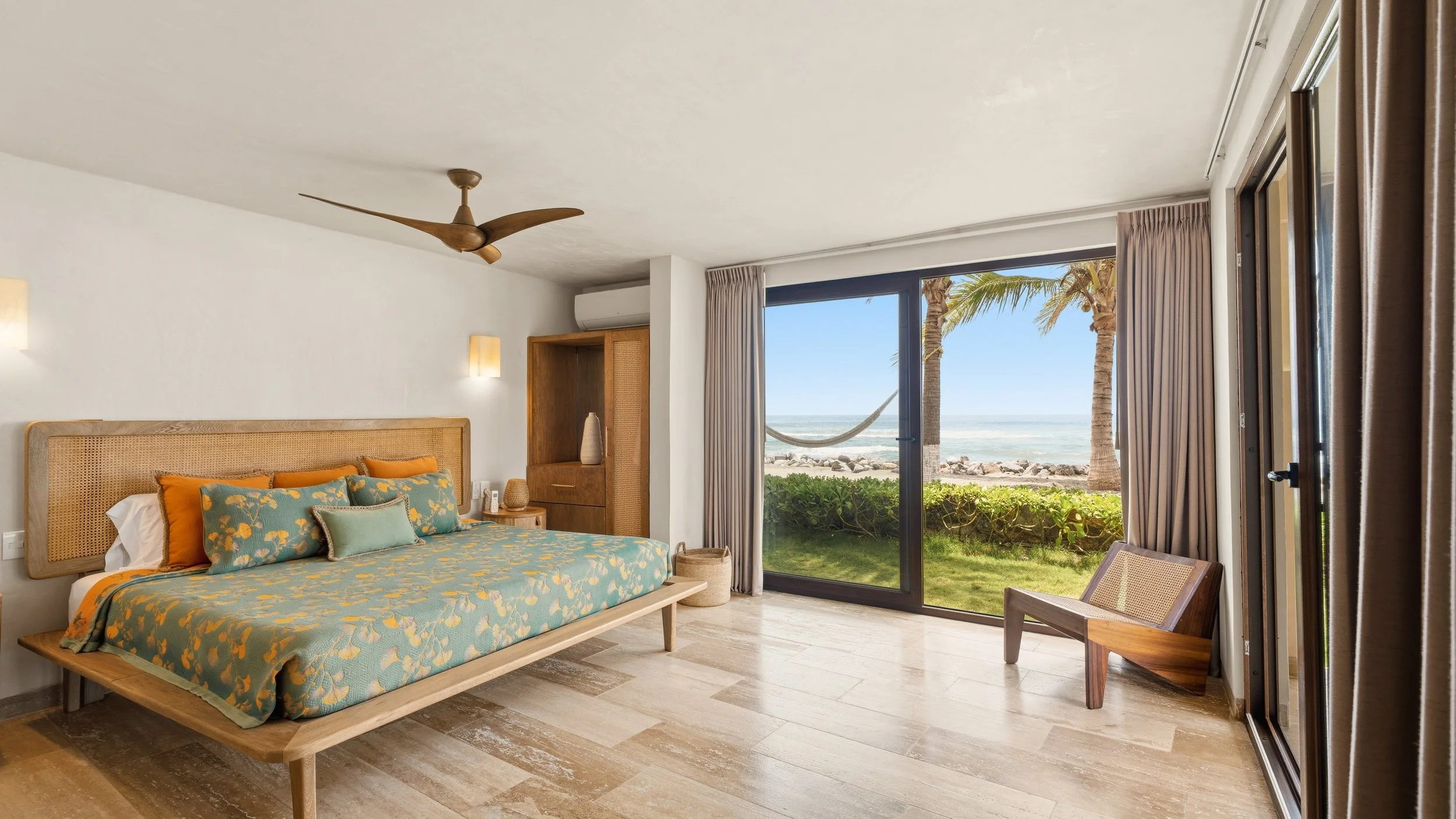 A bedroom with a sliding glass door leading to a beach view, a bed with colorful bedding, and tropical decor.