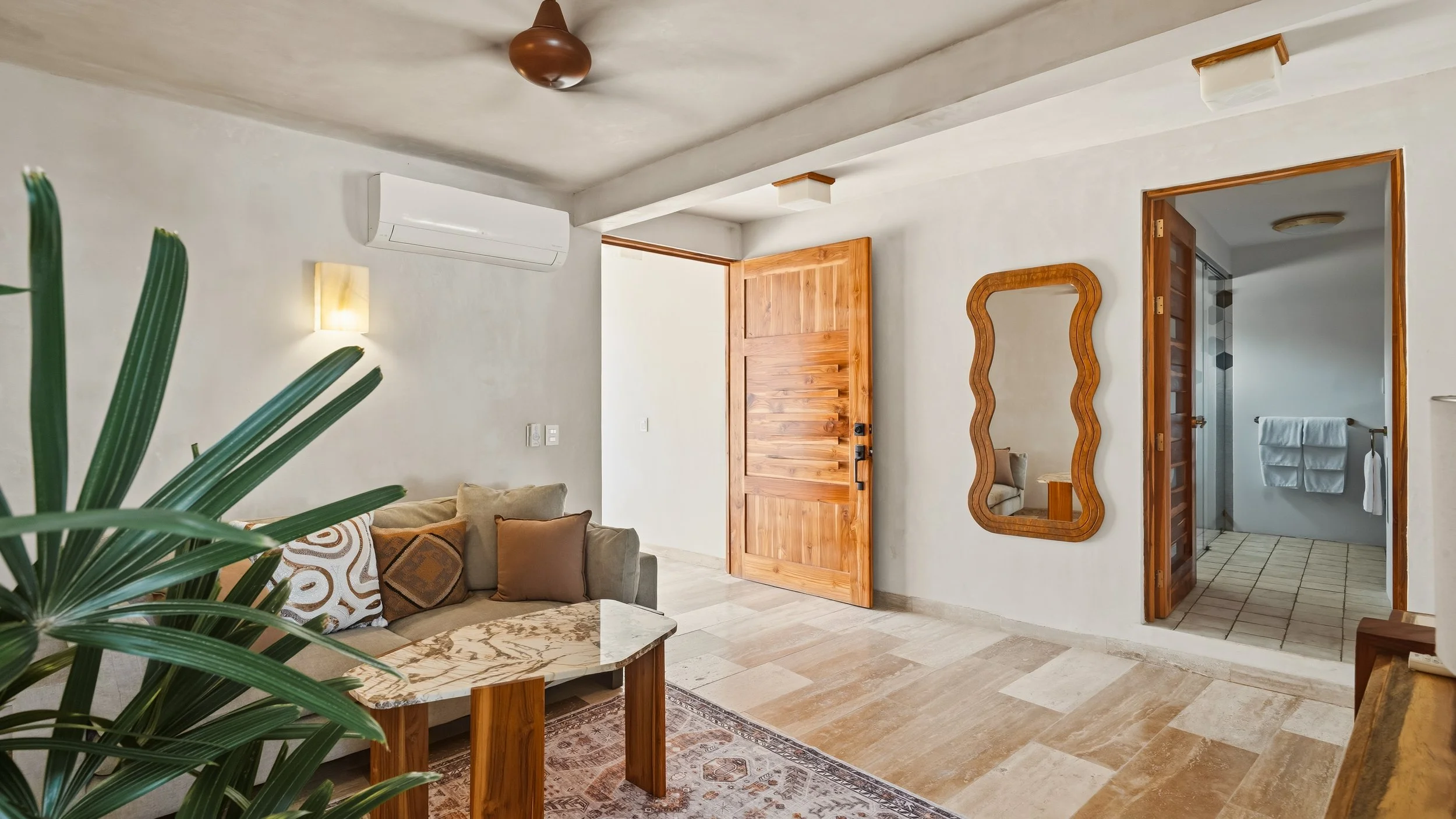 A cozy living room with white walls, a beige couch with patterned and solid pillows, a marble-top coffee table, a full-length wooden mirror, a wooden door, an air conditioning unit, and a doorway leading to a bathroom with white towels.