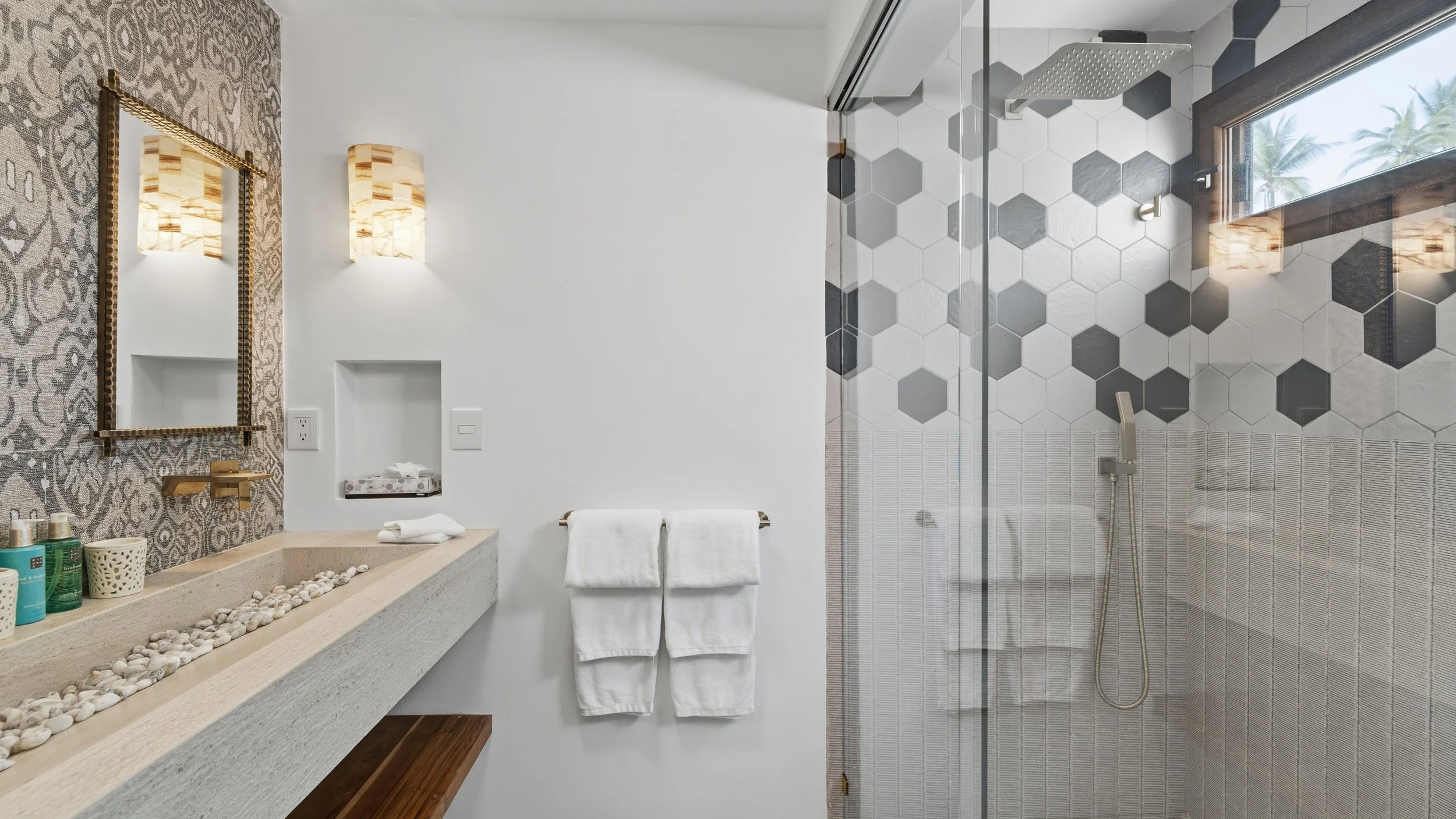 Modern bathroom with a vanity, mirror, towel rack, and walk-in shower with black and white hexagonal tiles and a large window with palm trees outside.