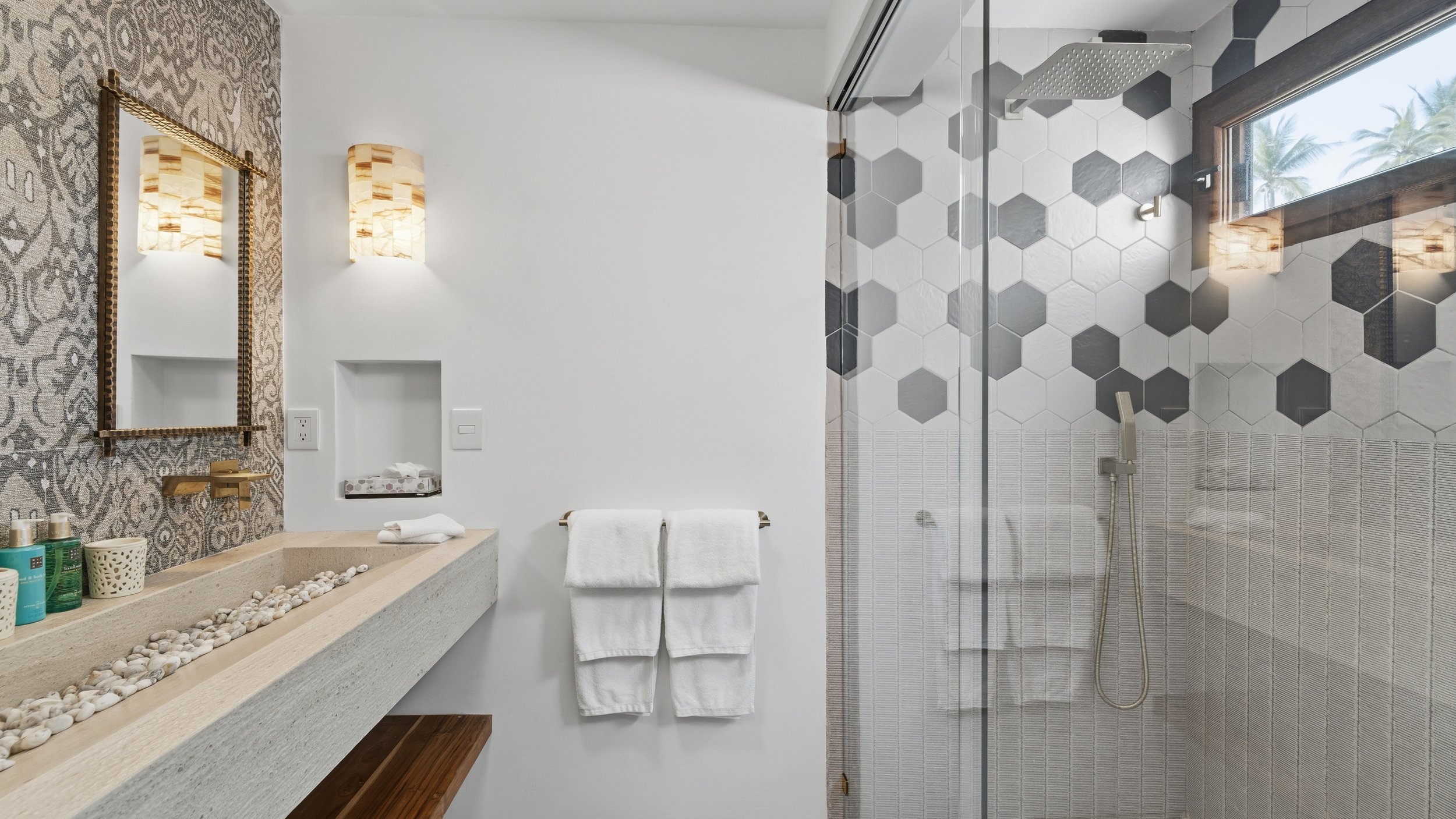Modern bathroom with a beige concrete sink, salt stones, towels, a mirror, decorative wall lights, and a walk-in shower with black and white hexagon tiles, large window with palm trees outside.