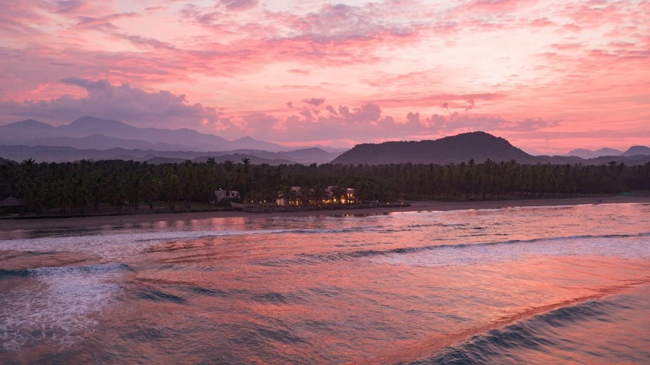 Sunset over the ocean, with pink and purple clouds, distant mountains, and palm trees along the shoreline.
