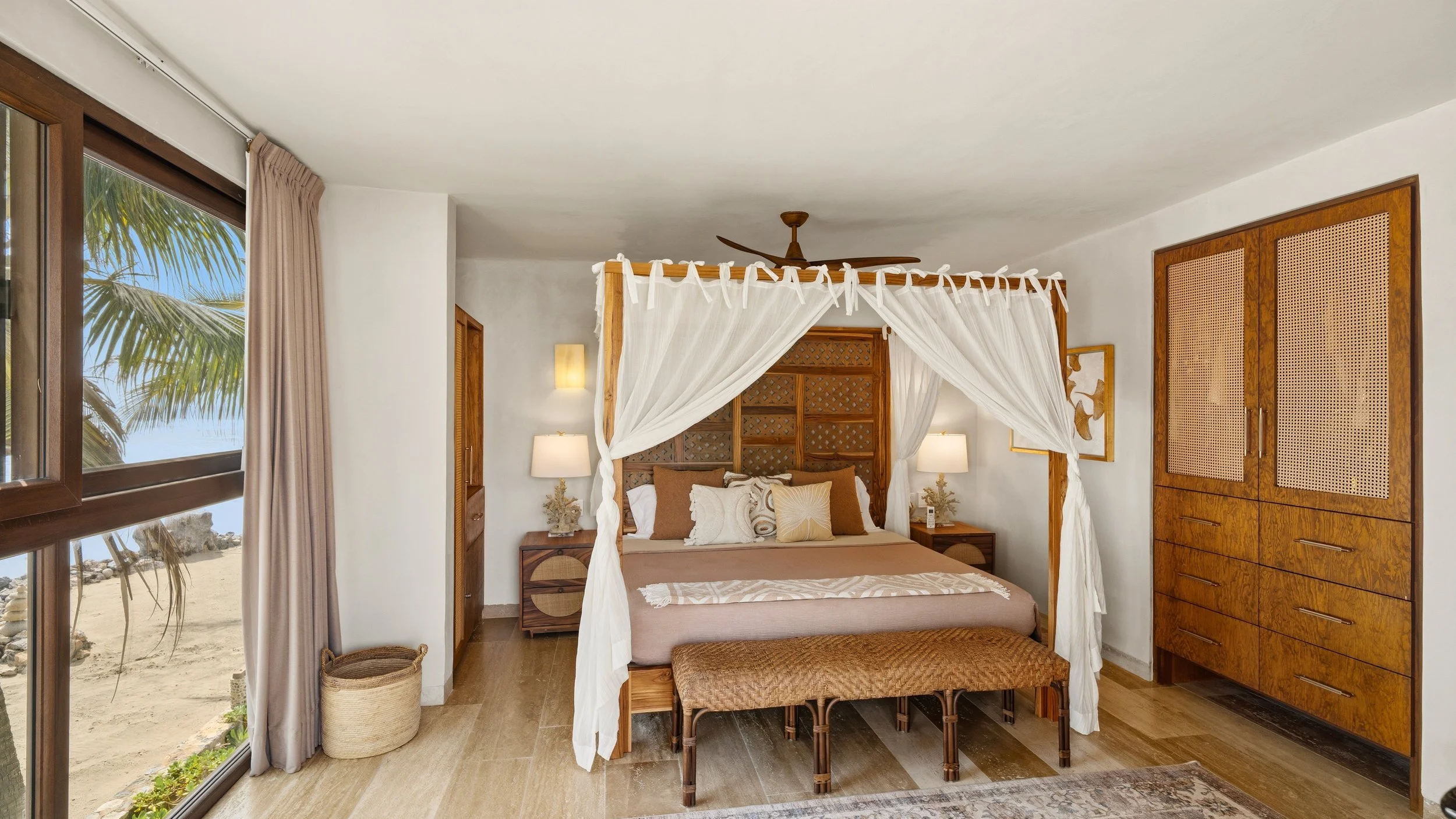 Bedroom with a four-poster bed, nightstands, lamps, wooden cabinets, and a window showing a beach scene with palm trees.