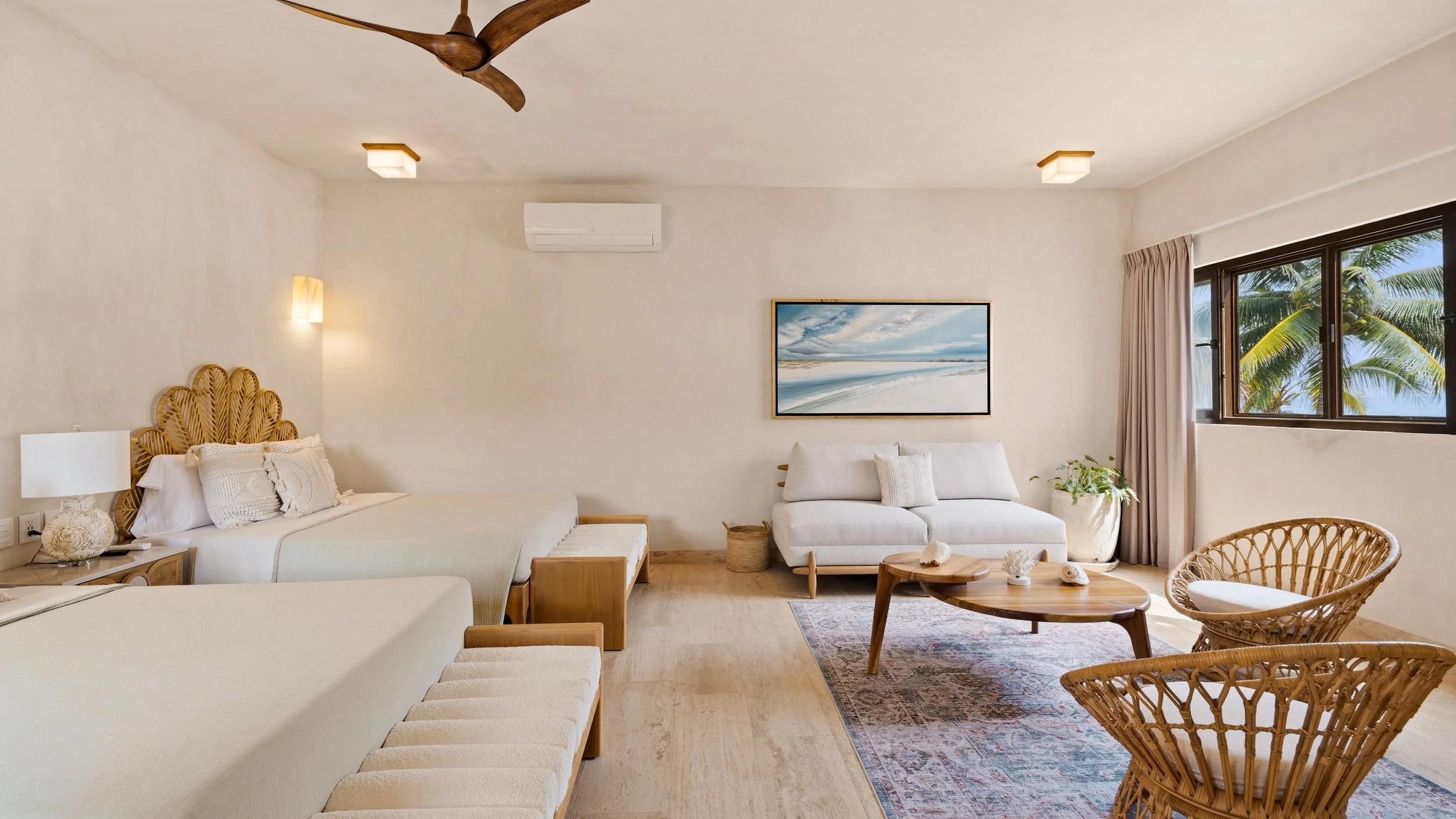 Bright and airy living room with white walls, a white sofa with textured pillows, natural wood furniture, a large window showing palm trees outside, and a coastal painting on the wall.