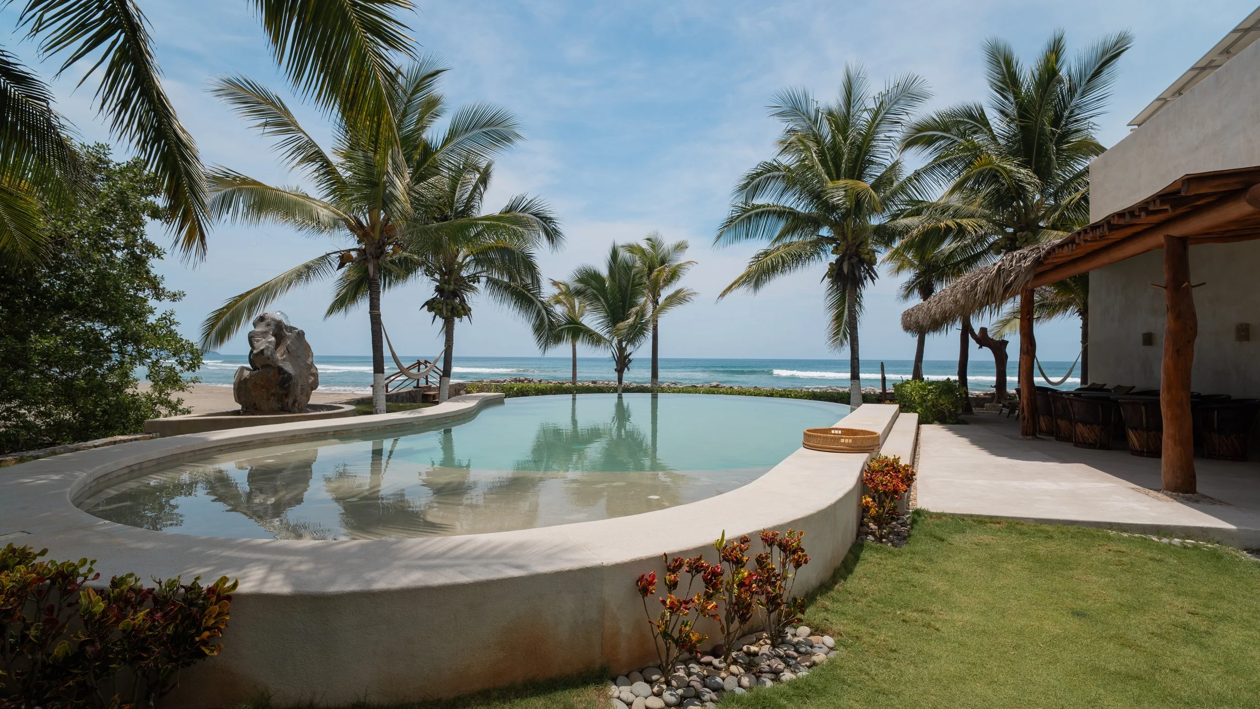 A backyard view of a small pool overlooking the ocean with tall palm trees, a hammock, a wicker basket, and a thatched-roof patio area.