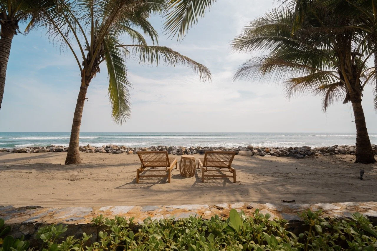 Two wooden lounge chairs with cushions facing the ocean on a sandy beach, flanked by two palm trees, with a small table between them and a rock barrier in the background.
