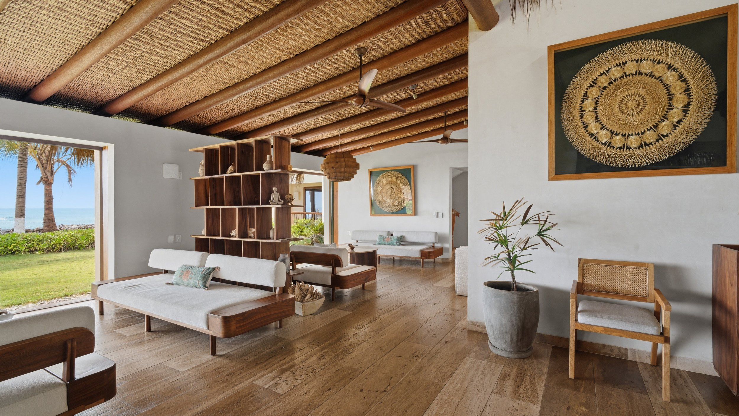 Living room with white sofas, wooden chairs, a potted plant, wall art, and open view of a garden and ocean with palm trees.