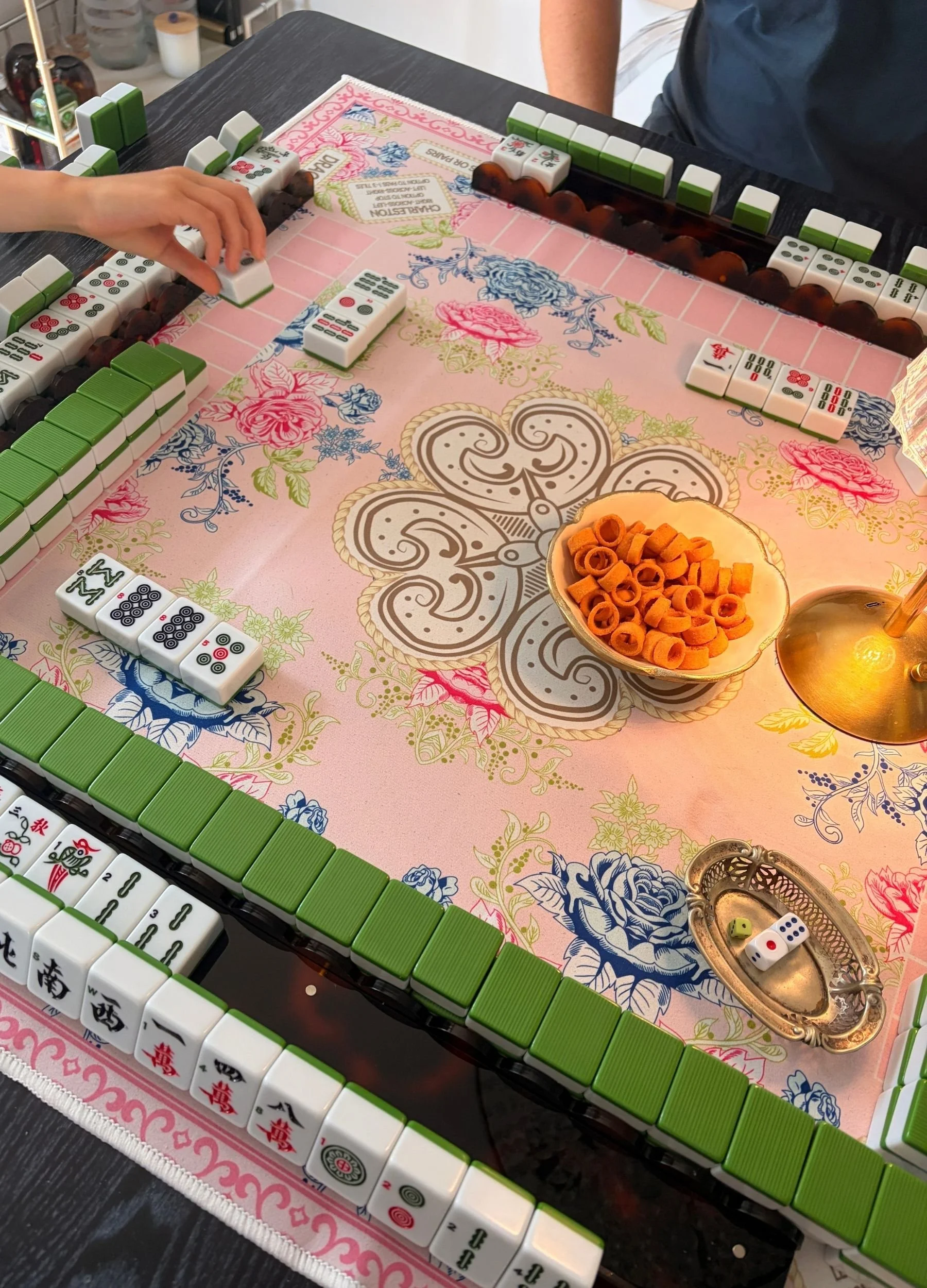 Mahjong game night setup with green tiles, pink floral mat, tile racks and snacks on the table.