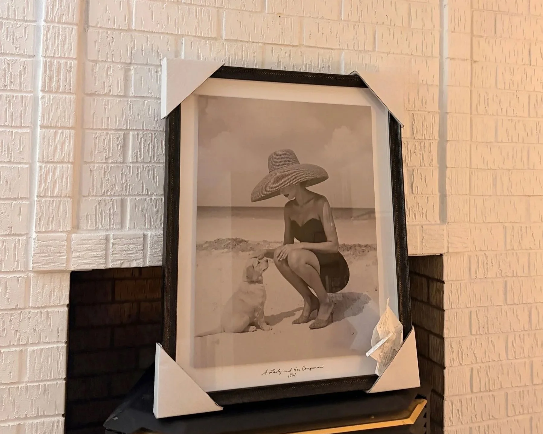 A vintage-style black and white framed print resting above the black fireplace against the painted white brick wall.