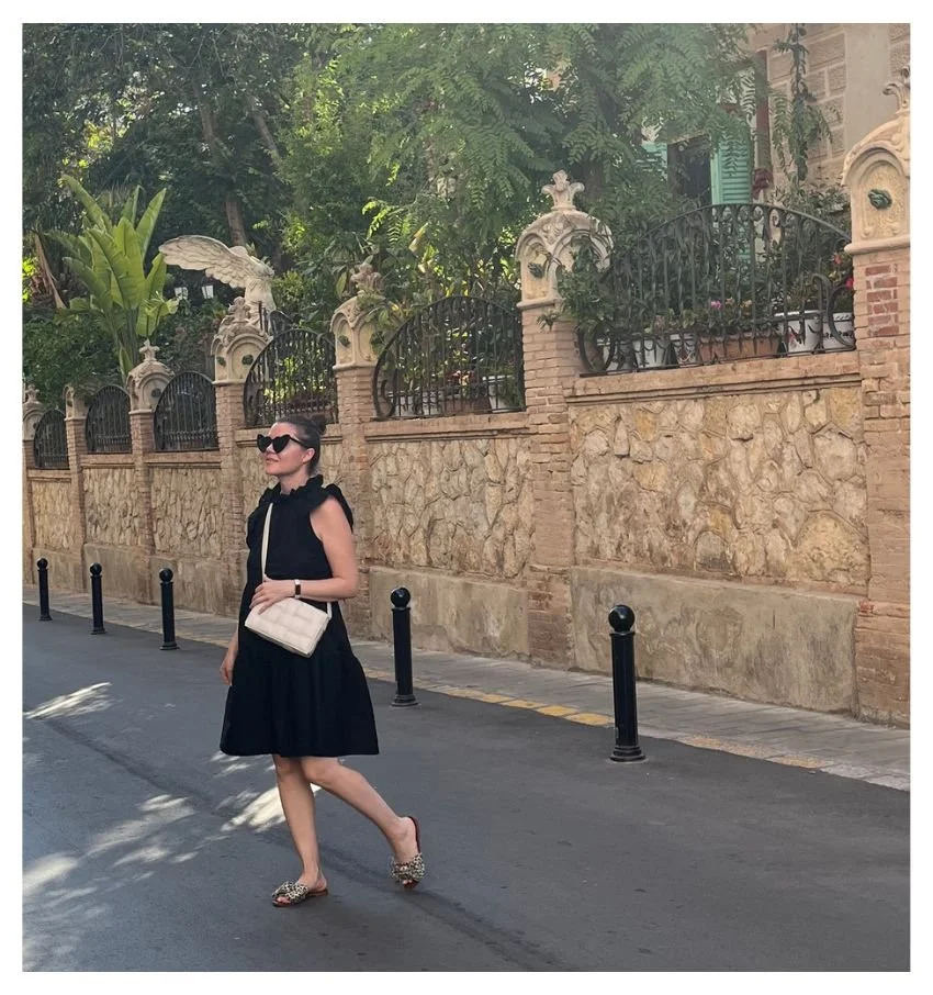 Woman in a black outfit walking past an ornate garden entrance in Sitges, Spain.