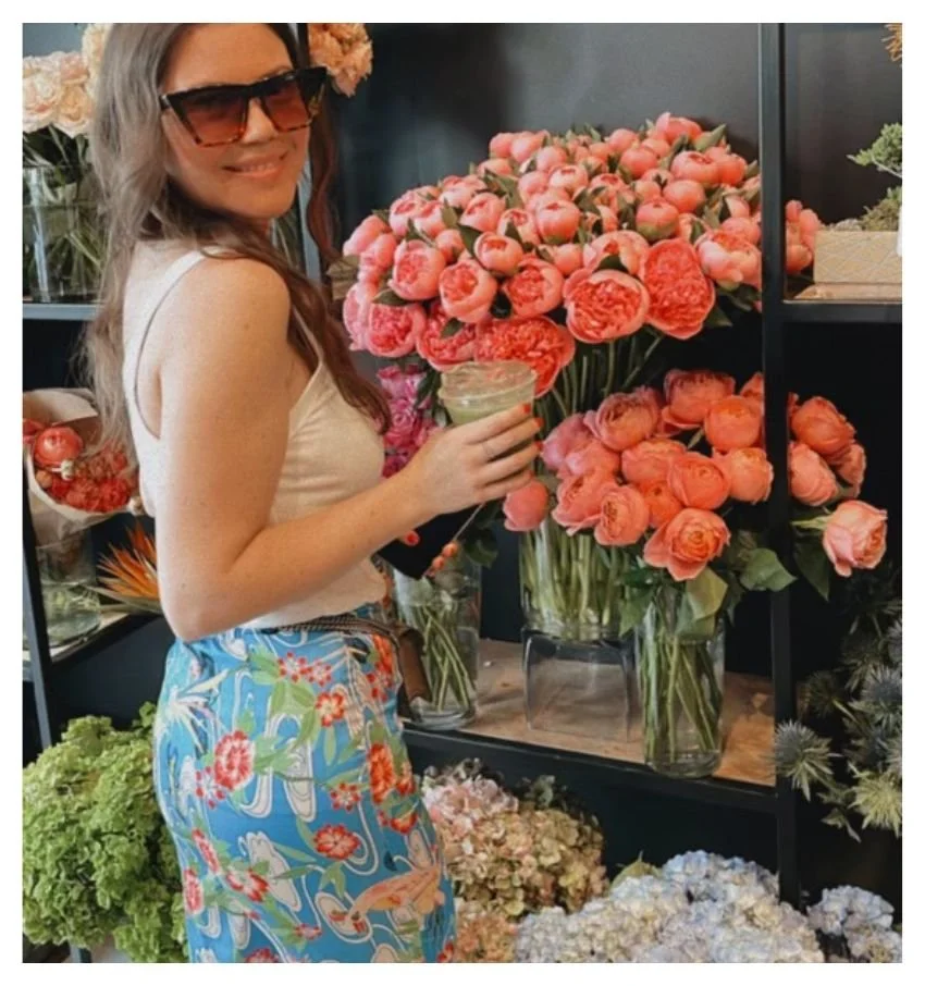 Woman standing in a flower shop surrounded by coral roses and colorful blooms.