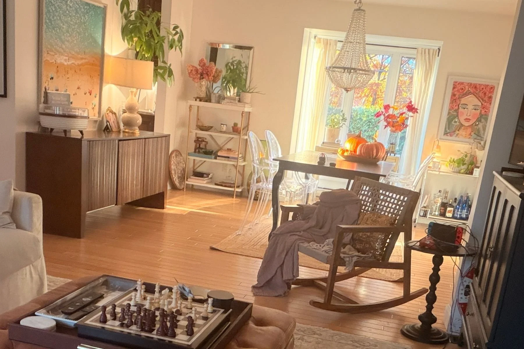 Cozy sunlit living and dining room with warm wood furniture, plants and autumn decor.